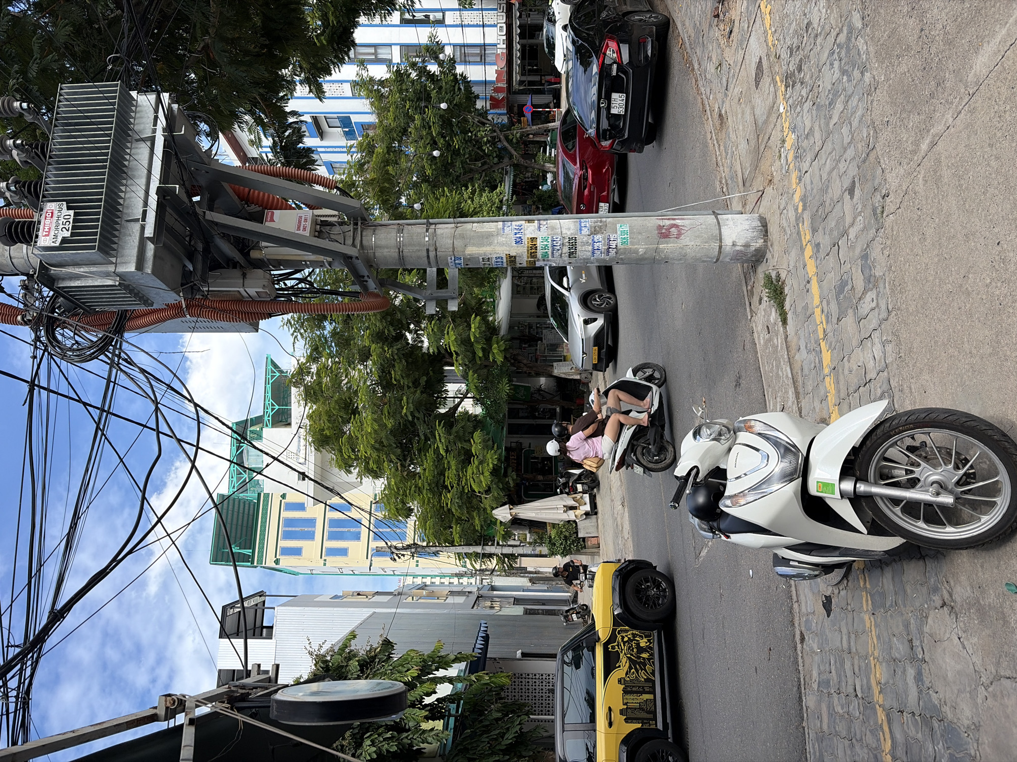 The image depicts a street scene in Da Nang, Vietnam, with a focus on an urban environment characterized by its utility infrastructure and traffic. Dominating the center of the frame is a large electrical transformer mounted on a utility pole, adorned with numerous wires and orange conduit. The pole itself is covered in various stickers and notices.
In the foreground, a white Honda scooter is parked on the sidewalk. The street is asphalt, marked with yellow lines and bordered by a tiled sidewalk. Further down the street, a yellow and black SUV with decorative graphics is parked. Two individuals are riding a scooter, a woman in a pink shirt and white shorts, and a man behind her, both wearing helmets.
The background features multi-story buildings, some with modern architecture and blue accents, interspersed with trees. The sky is partly cloudy. The overall scene suggests a typical day in a bustling Vietnamese city, with everyday activities such as commuting by scooter and the presence of essential urban services.