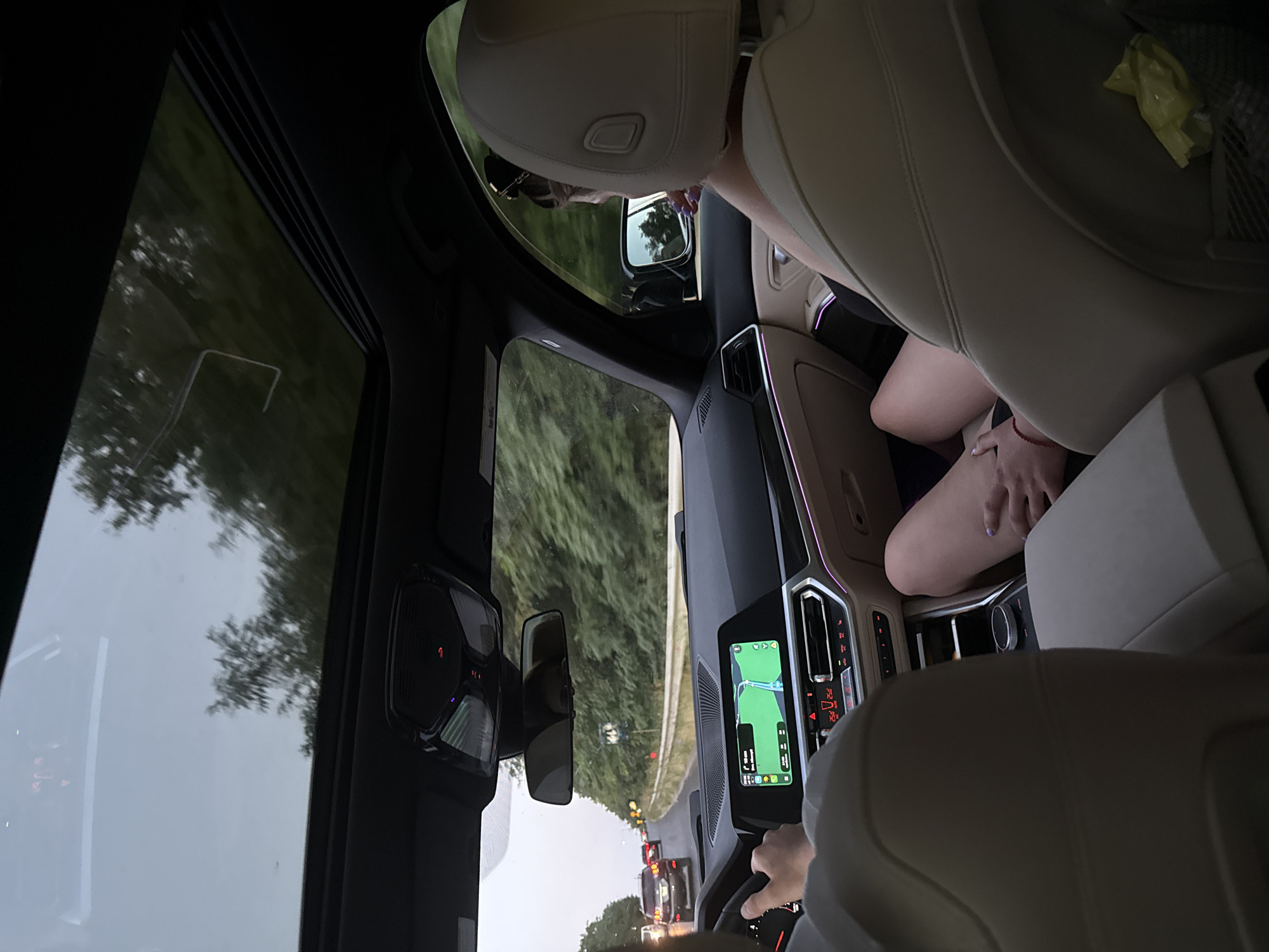 The image shows the interior of a car, looking forward from the back seat. A person's hands are visible on the steering wheel, indicating they are driving. The dashboard features a large infotainment screen displaying a navigation map, and climate controls with red temperature readouts. A sunroof is open, revealing a cloudy sky and trees. In the passenger seat, a person's legs and arm are visible, resting on the center console. Outside the car, other vehicles are seen on a road lined with dense green trees, suggesting a rural or suburban setting. The overall impression is a journey through a landscape. The location is identified as Fundul Galbenei, Moldova.