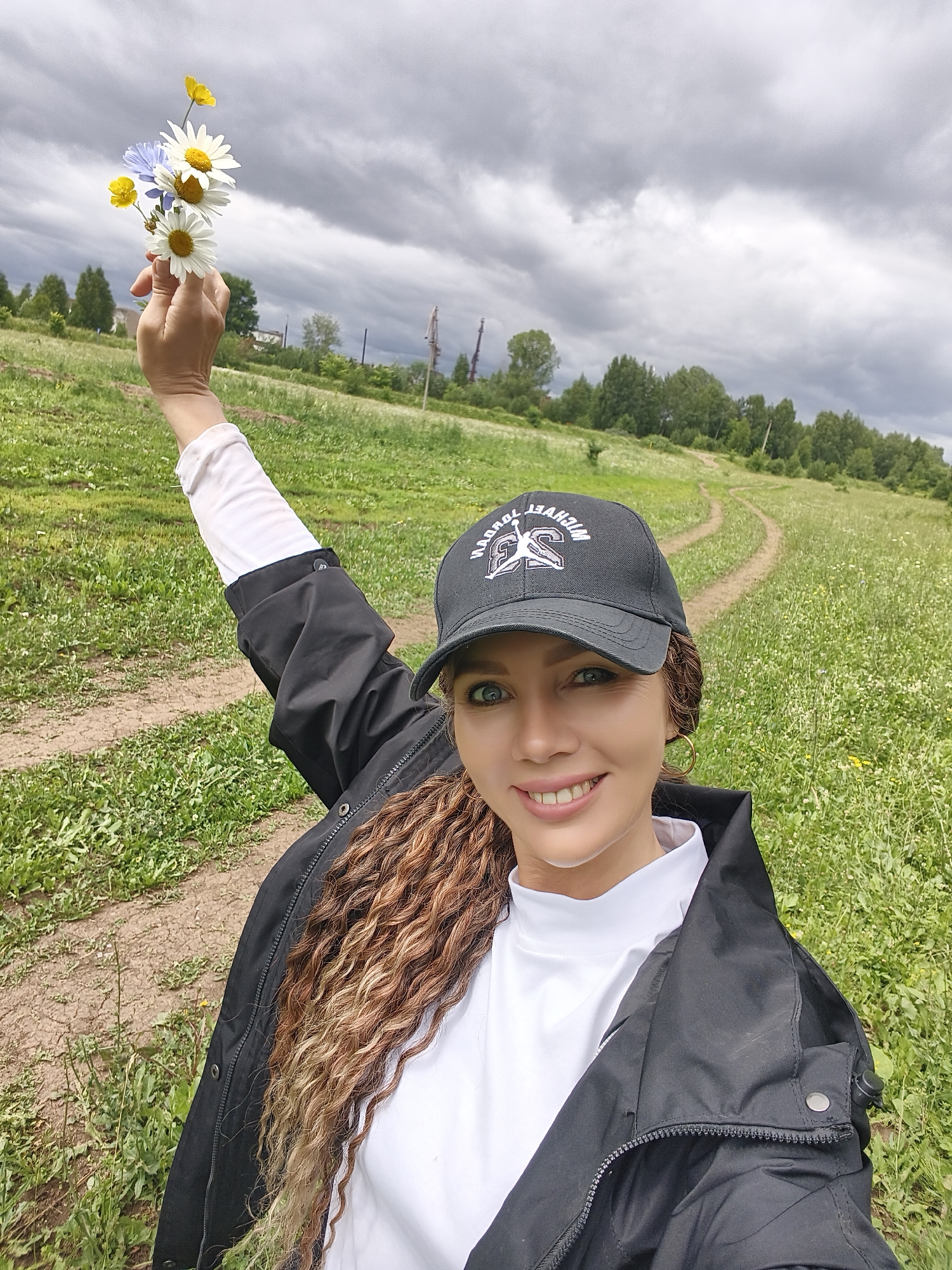 A woman stands in a grassy field, holding a small bouquet of daisies, buttercups, and blue wildflowers. She is wearing a black baseball cap with a white logo that appears to say "Michael Jordan" with a basketball player silhouette. She also has on a white turtleneck and a black jacket. The woman has long, curly brown hair and is smiling at the camera.
The background shows a dirt path winding through the field and a line of trees in the distance under a cloudy, overcast sky. The scene suggests a rural or natural setting, possibly a park or countryside. The weather appears to be cool and perhaps threatening rain, given the dark clouds. The overall mood is cheerful and relaxed, with the woman's smile and the bright wildflowers contributing to a pleasant atmosphere. No specific time of day is indicated, but the lighting suggests it is daytime. The location context is Noviy, Russia.