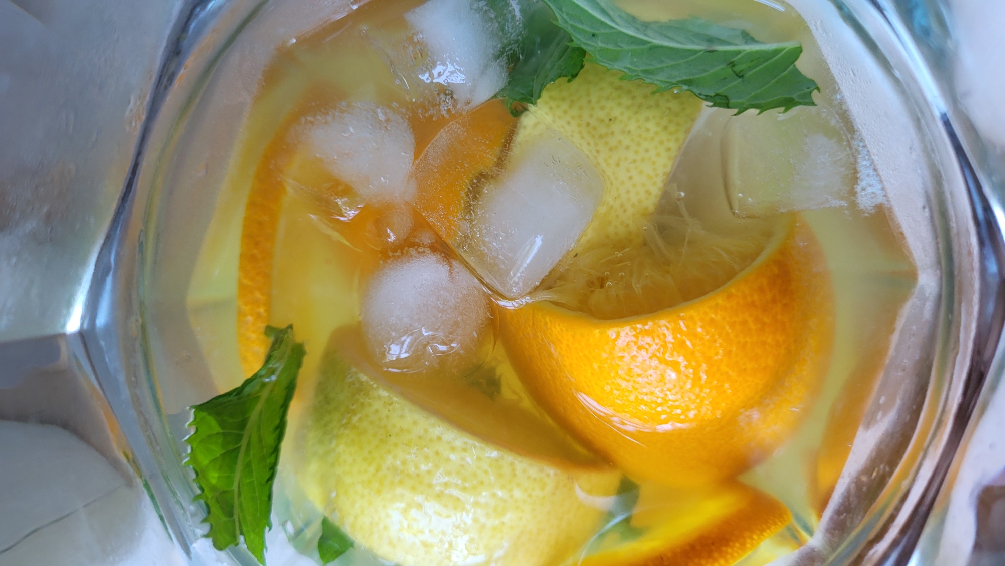 This is a close-up, top-down shot of a clear glass pitcher filled with a refreshing beverage. The drink is a pale yellow, suggesting it is some form of lemonade or citrus-infused water. Visible within the pitcher are several ice cubes, contributing to the chilled appearance of the drink.
Submerged in the liquid are slices and wedges of citrus fruits, including oranges and lemons. The oranges exhibit their characteristic vibrant orange peel, while the lemons are a bright yellow. The fruits appear fresh, adding to the overall visual appeal of a cool, summery drink. Interspersed among the ice and fruit are sprigs of fresh mint, their green leaves adding a touch of color and indicating a hint of mint flavor in the beverage.
The lighting in the image is bright, suggesting it was taken during the daytime, possibly outdoors or near a window. The reflection of light on the glass and the condensation on the pitcher's surface further enhance the impression of a cold and thirst-quenching drink. The focus is sharp on the contents of the pitcher, highlighting the textures of the ice, fruit, and mint. There is no visible text in the image. The scene evokes a sense of refreshment and relaxation, perfect for a warm day.