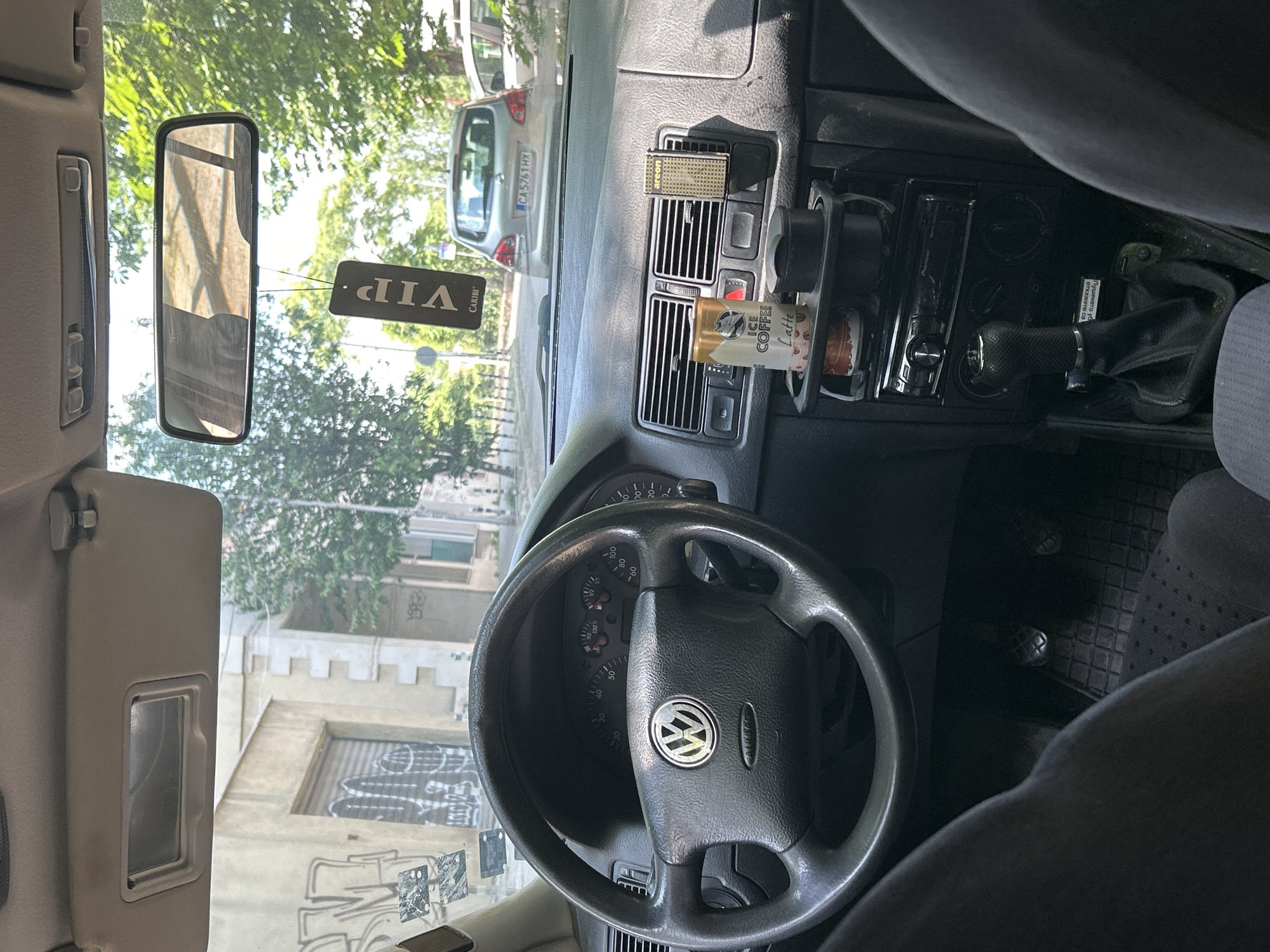 The image depicts the interior of a Volkswagen Golf Mk4 from the driver's perspective, looking forward onto a city street. The black steering wheel, centered in the foreground, features the Volkswagen logo and "AIRBAG" lettering. Behind the steering wheel, the instrument cluster shows the speedometer needle at zero and the engine temperature gauge at approximately 90 degrees Celsius.
Above, a beige sun visor with a rectangular vanity mirror is extended. A black "VIP CARIBI" air freshener hangs from the rearview mirror, which reflects a blurred interior view.
The dashboard contains two central air vents. An "areon" branded air freshener is clipped into the right vent. A "Ice Coffee Latte" can rests in a black plastic cup holder system mounted below the vents. Below the cup holder is a Pioneer brand car stereo unit. The black manual gear shift lever is visible below the stereo, and a partially visible white sticker with Cyrillic text, including "Пътуването", is affixed nearby. The floor shows a black textured rubber mat, and the clutch and brake pedals are partially visible in the driver's footwell.
Through the windshield, a street scene unfolds under bright daylight. A silver hatchback car with the Bulgarian license plate "CA 5761 HX" is parked directly ahead, confirming the location as Sofia, Bulgaria. Another white vehicle is partially visible further down the street. The left side of the street is lined with multi-story buildings, some displaying extensive graffiti, particularly on a closed garage door. Green trees line the right side of the street. No individuals are visible in the scene.