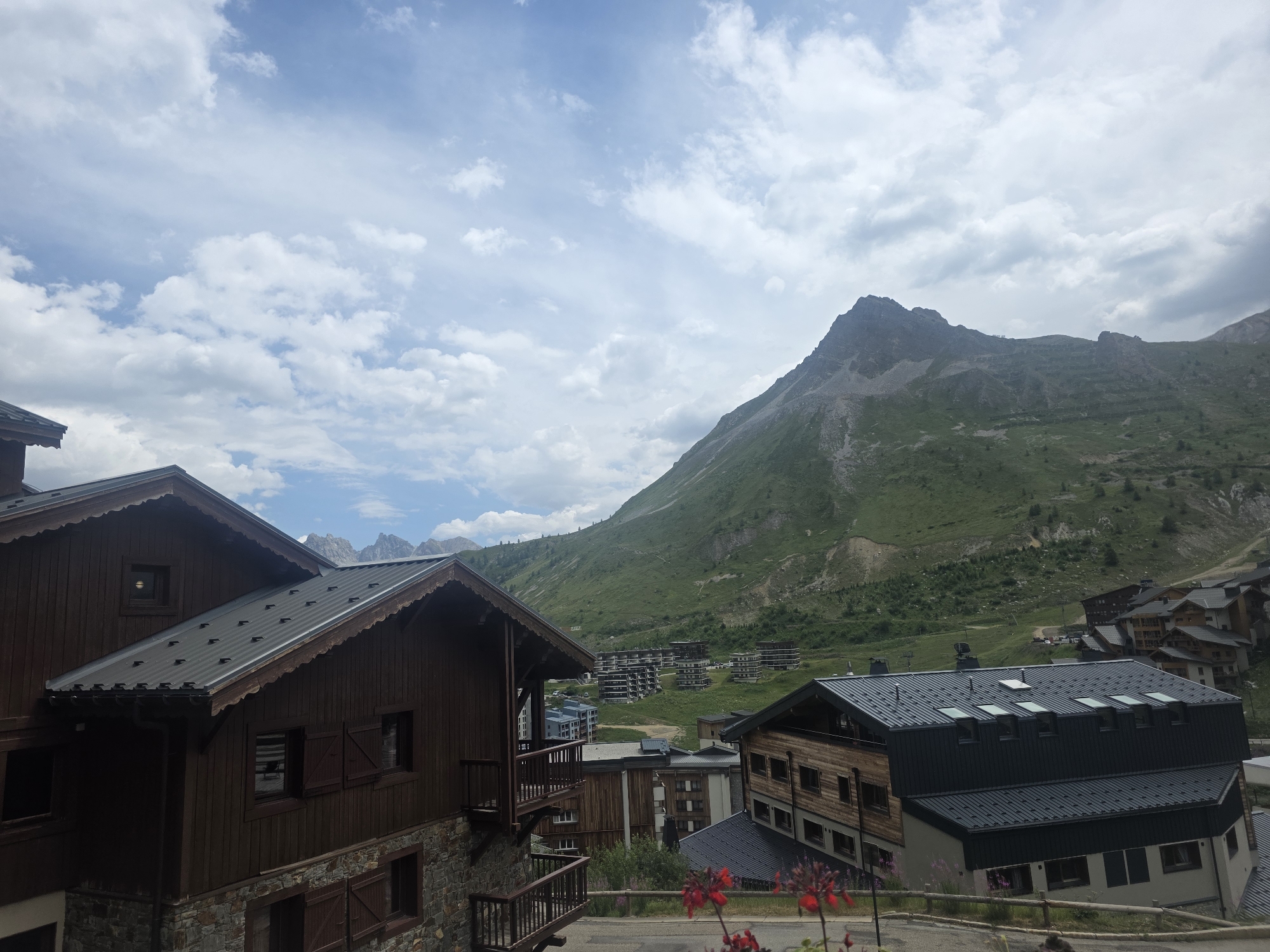 The image captures a view of the Val Claret sector of Tignes, France, a mountain resort town. In the foreground, traditional wooden chalets with stone foundations and dark metal roofs are visible. These buildings feature balconies and shutters. Beyond the chalets, a cluster of modern, multi-story apartment buildings, typical of ski resorts, are nestled in the valley. The background is dominated by a large, verdant mountain slope, characterized by its steep incline and scattered vegetation. The sky above is filled with a mix of white and gray clouds against a pale blue backdrop. Red flowers are blooming in the lower right corner of the frame, adding a touch of color to the scene. The overall impression is one of a summer day in a mountain resort, with a blend of traditional and contemporary architecture set against a dramatic natural landscape.