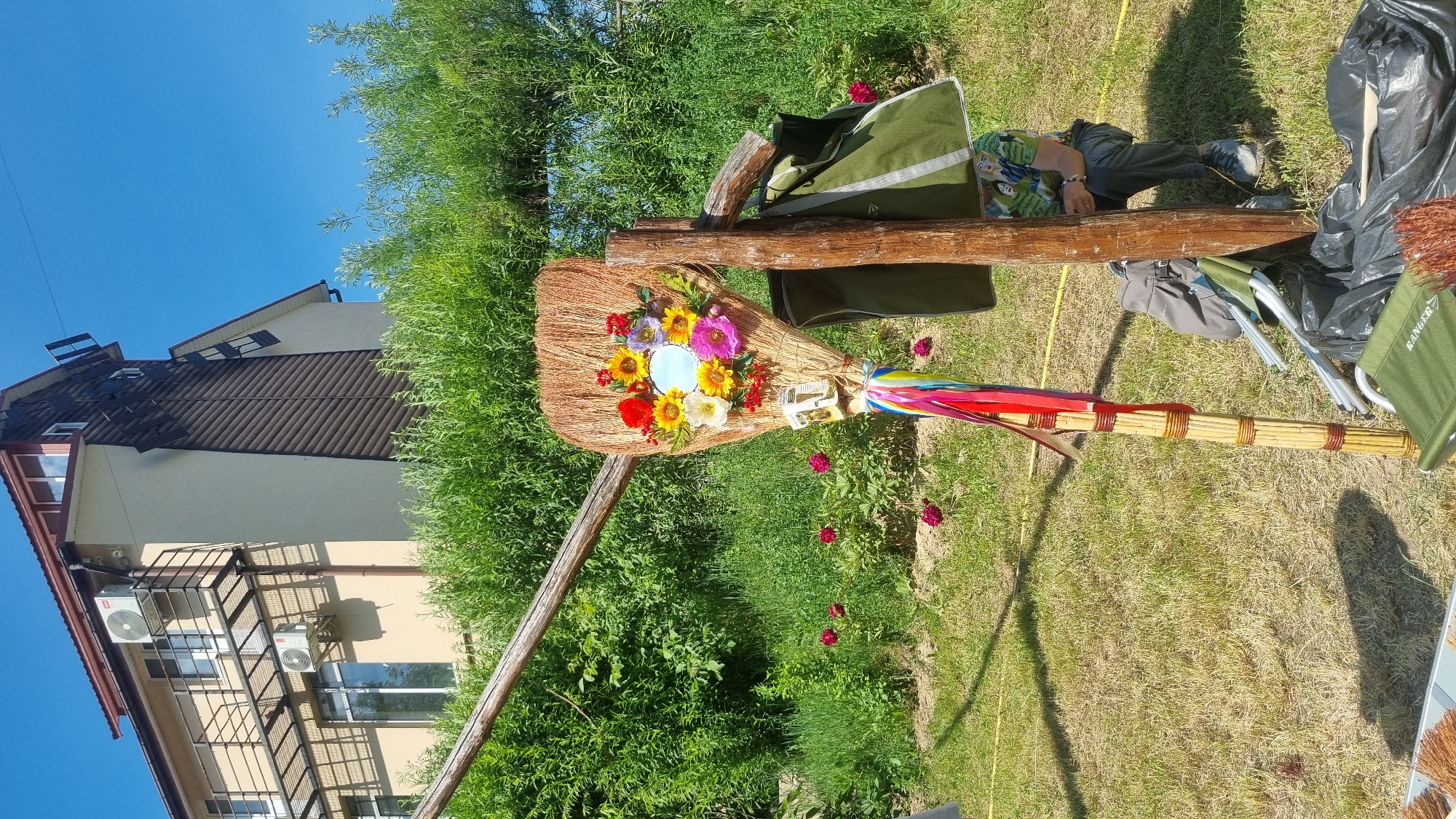 The image displays an outdoor scene in Melnyky, Ukraine, under a clear, cloudless blue sky on a bright, sunny day.
In the foreground, a decorative broom stands upright, leaning against a rustic wooden post. The broom's head, composed of light-colored natural fibers, is adorned with a central, small, round mirror surrounded by an arrangement of artificial flowers in various colors, including red, yellow, purple, pink, and white. Below the flower arrangement, two small, rectangular white objects with visible text or design are affixed. The broom handle, constructed from sections of light-colored wood or bamboo, is wrapped with darker material, and colorful ribbons (red, blue, yellow, green, and white) are tied around its upper part, hanging downwards.
To the immediate right of the broom structure, a dark green fabric bag with a lighter stripe hangs from a horizontal wooden beam. Partially obscured by the vertical wooden post and the hanging bag, a person is seated on a small, dark green folding chair. The individual wears dark trousers, grey athletic shoes, and a light green short-sleeved t-shirt with colorful printed graphics. On the ground next to the person are a grey backpack, a partially folded green chair, and a black plastic bag containing additional items, including what appears to be more broom heads. A yellow hose or cable traverses the lower-middle foreground. Several vibrant pink or red flowers are blooming on the ground behind the broom.
The mid-ground is dominated by dense, lush green foliage, consisting of bushes and possibly small trees. A large wooden beam extends diagonally from the lower-left corner towards the upper-center, forming part of a larger wooden support structure.
In the background, a multi-story residential building with a reddish-brown pitched roof is visible. The building's exterior is primarily light beige or cream, with a distinct vertical section on the right side clad in dark brown horizontal siding. A metal balcony with a grid-patterned floor extends from the left side of the building, and two outdoor air conditioning units are mounted on the wall below it. A dark, slatted, ladder-like structure is affixed vertically to the brown siding on the far right side of the building, extending towards the roofline.