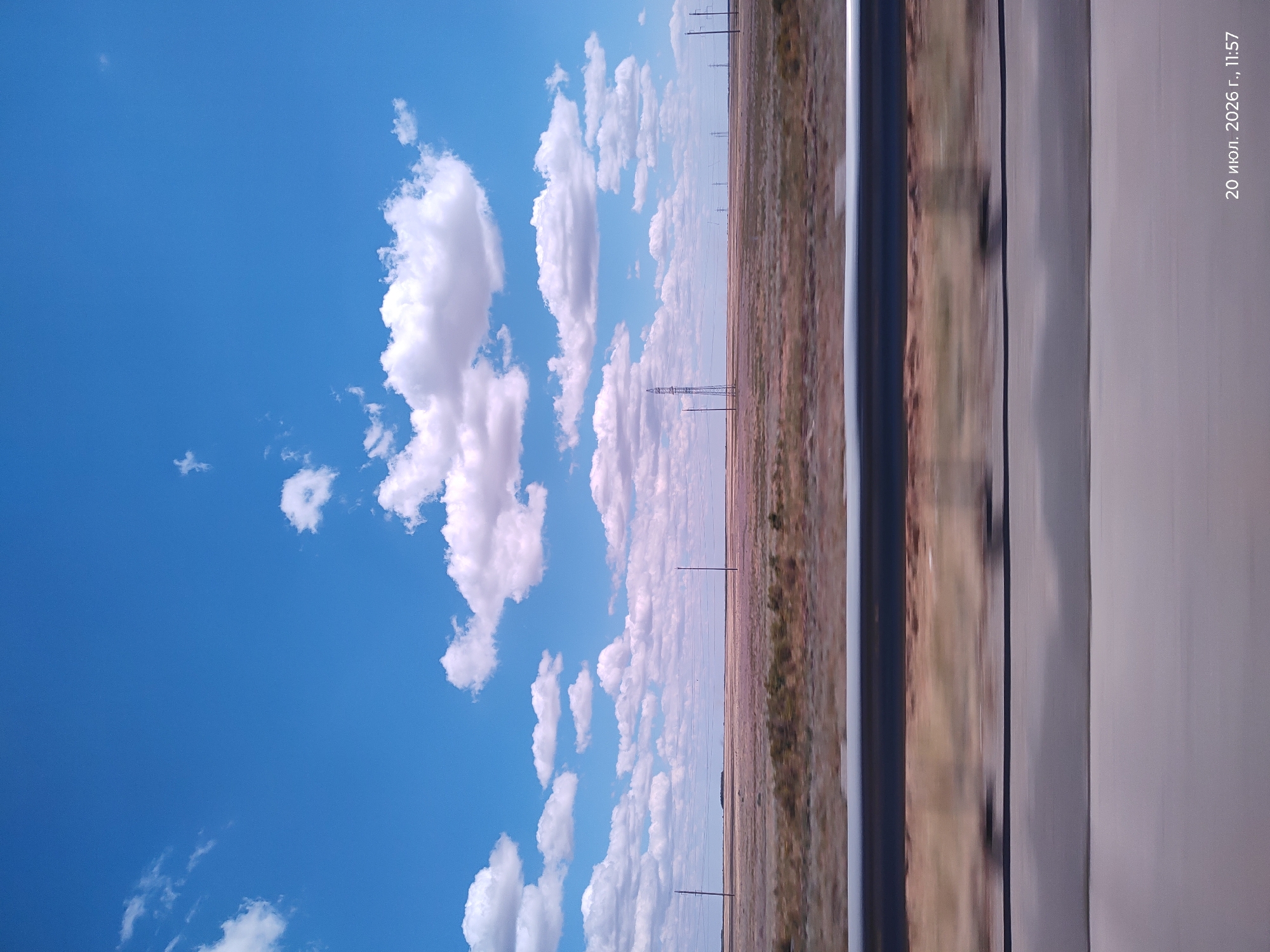 The image captures a vast, flat landscape under a bright blue sky with scattered white clouds, characteristic of the steppes near Dams, Kazakhstan. The foreground is dominated by a blurred representation of a road or ground, suggesting movement. A metallic guardrail runs horizontally across the mid-ground, separating the road from the arid terrain. Beyond the guardrail, the landscape unfolds as a dry, brown plain dotted with sparse vegetation and a few scattered power poles. In the distance, several tall power pylons are visible, indicating infrastructure within this remote area. The sky is a vibrant blue, with puffy white clouds creating a dynamic overhead view. The timestamp in the bottom right corner reads "20 июля 2026 г., 11:57", indicating the date and time the photograph was taken.