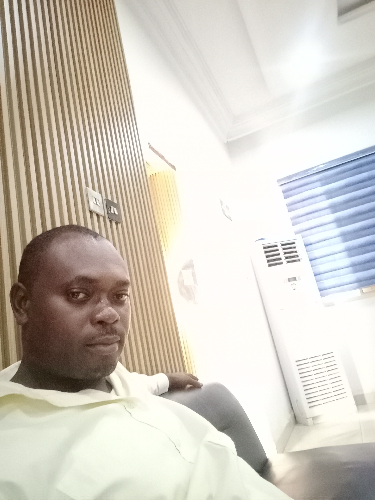 The image shows a man in an interior setting, seemingly in Gwarinpa, Nigeria. The man is wearing a light yellow shirt and is seated. Behind him is a decorative wall feature with vertical wooden strips, along with light switches. Further back, there's an air conditioning unit standing near a window with blue blinds. The ceiling is white and includes recessed lighting. The man is the primary subject and appears to be posing for the picture.