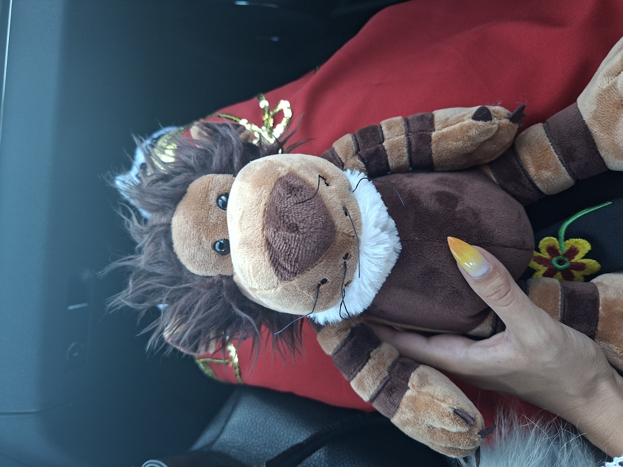A stuffed lion toy is held by a person's hand in what appears to be the interior of a vehicle. The lion is brown with lighter brown stripes on its arms and legs, and a fluffy mane. It has a white collar and a brown nose with black whiskers. The person's hand has a long, pointed fingernail painted with a gradient of yellow to white. A red fabric with gold accents is visible behind the lion. The background is dark and out of focus, suggesting a car's interior. The overall scene is an indoor, artificial environment.