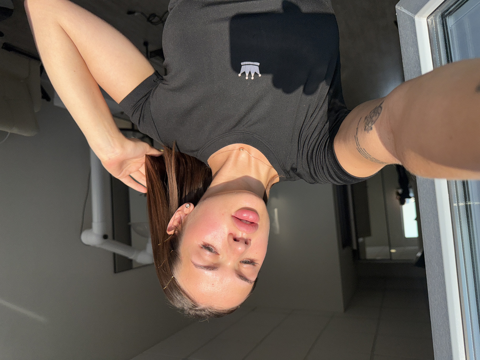A young woman with light brown hair and fair skin is pictured from the chest up, standing indoors. She is wearing a black t-shirt with a small, white crown emblem on the left side of the chest. A thin, rose-gold necklace with a small, shimmering pendant is visible around her neck. She has a small, diamond stud earring in her left ear. Her right hand is raised and resting near her head, with her fingers touching her hair. She appears to be looking downwards and to her left. Sunlight is casting a warm glow on her face and arm. Her left arm is extended forward, and a tattoo is visible on her upper arm. The background is out of focus but appears to be an office or indoor space with a hallway visible to the left and a chair in the distance to the right. The lighting suggests it is daytime.