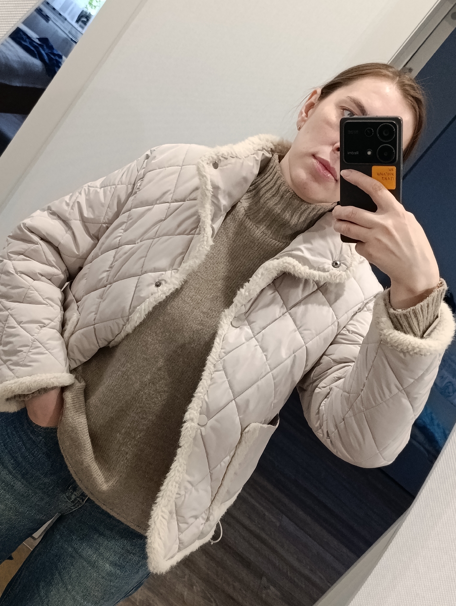A woman is taking a selfie in what appears to be a room with a reflective surface, possibly a mirror. She is wearing a tan quilted jacket with fur trim, a tan turtleneck sweater, and blue jeans. She is holding a smartphone with a black case, obscuring part of her face. The background shows a glimpse of what might be a window with a view of a building or structure. Based on the metadata, this image was taken in Chaikovsky, Russia.