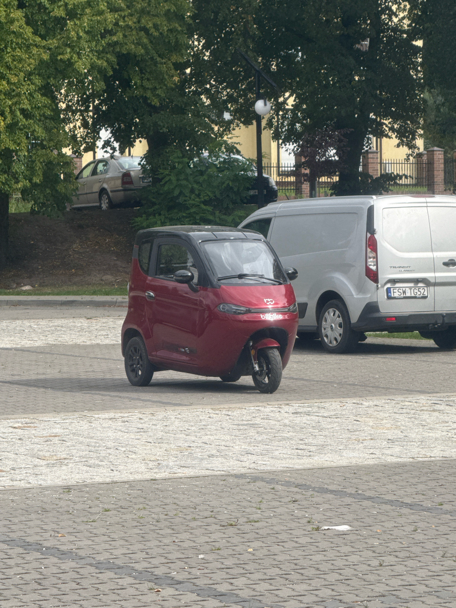 A red, three-wheeled microcar is parked on a paved lot, next to a silver van. The microcar has a black roof and doors. The van is a Ford Transit Connect, with the license plate FSW TG52. In the background, trees and a yellow building are visible. The scene is located in Łagów, Poland.