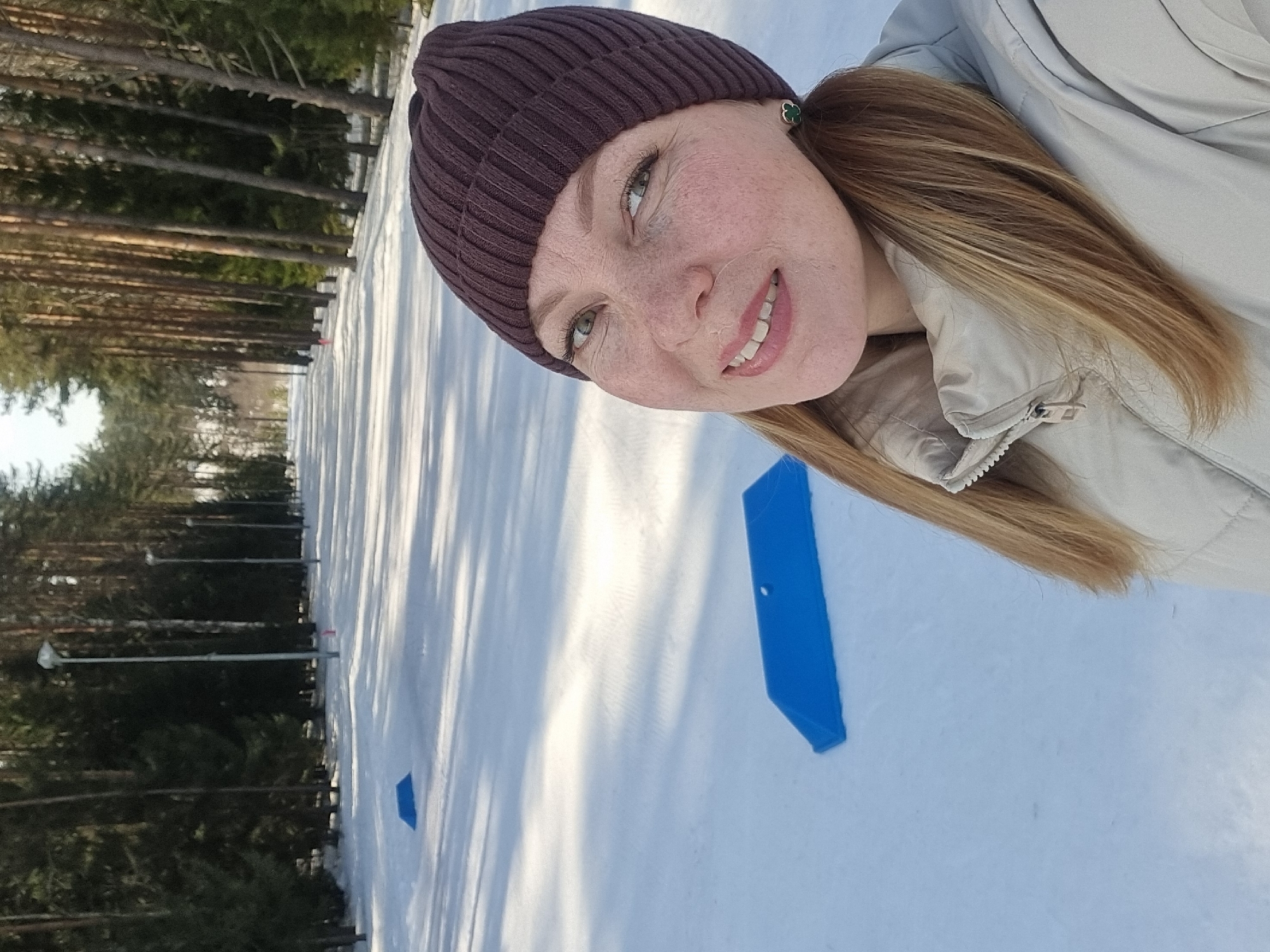The image depicts a female subject in a snowy outdoor setting, identified as Прикамский, Russia. The woman, with light brown hair, freckles, and green eyes, is wearing a dark brown ribbed knit beanie and a light beige puffer jacket. She is smiling and looking directly at the camera. The background features a wide, snow-covered path bordered by tall pine trees on both sides, with multiple streetlights visible along the path. Two blue, wedge-shaped objects are present on the snow in the foreground and midground. The scene is illuminated by natural light, casting long shadows across the snow.