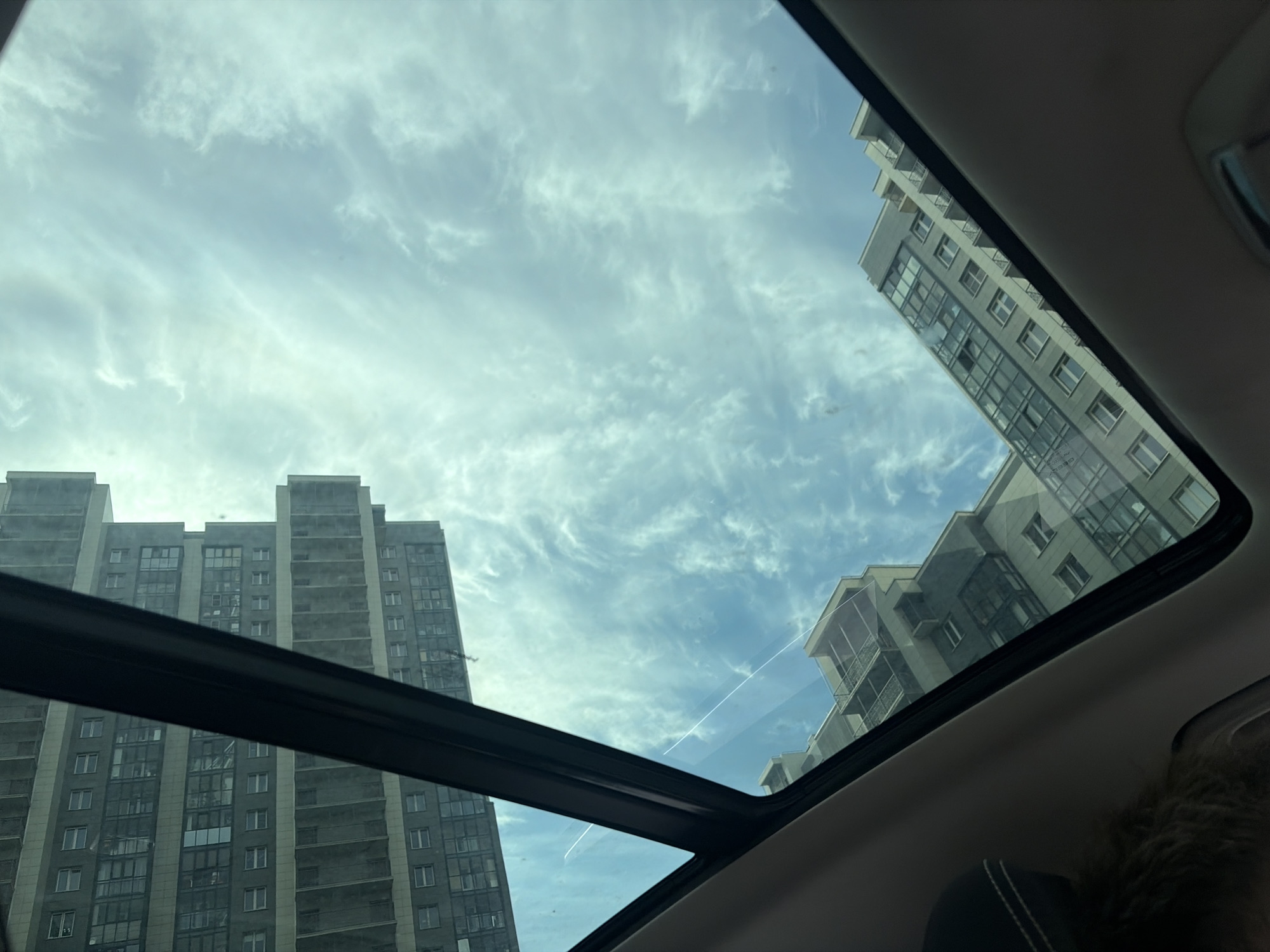 The image shows a view through a sunroof, likely in a car. The sky is visible with scattered clouds. Outside the sunroof, there are buildings, likely apartment buildings. The buildings are modern in design, with multiple floors and windows. The location is possibly Saint Petersburg, Russia, given the architectural style and the general appearance of the buildings. There are no people visible in the sunroof area.