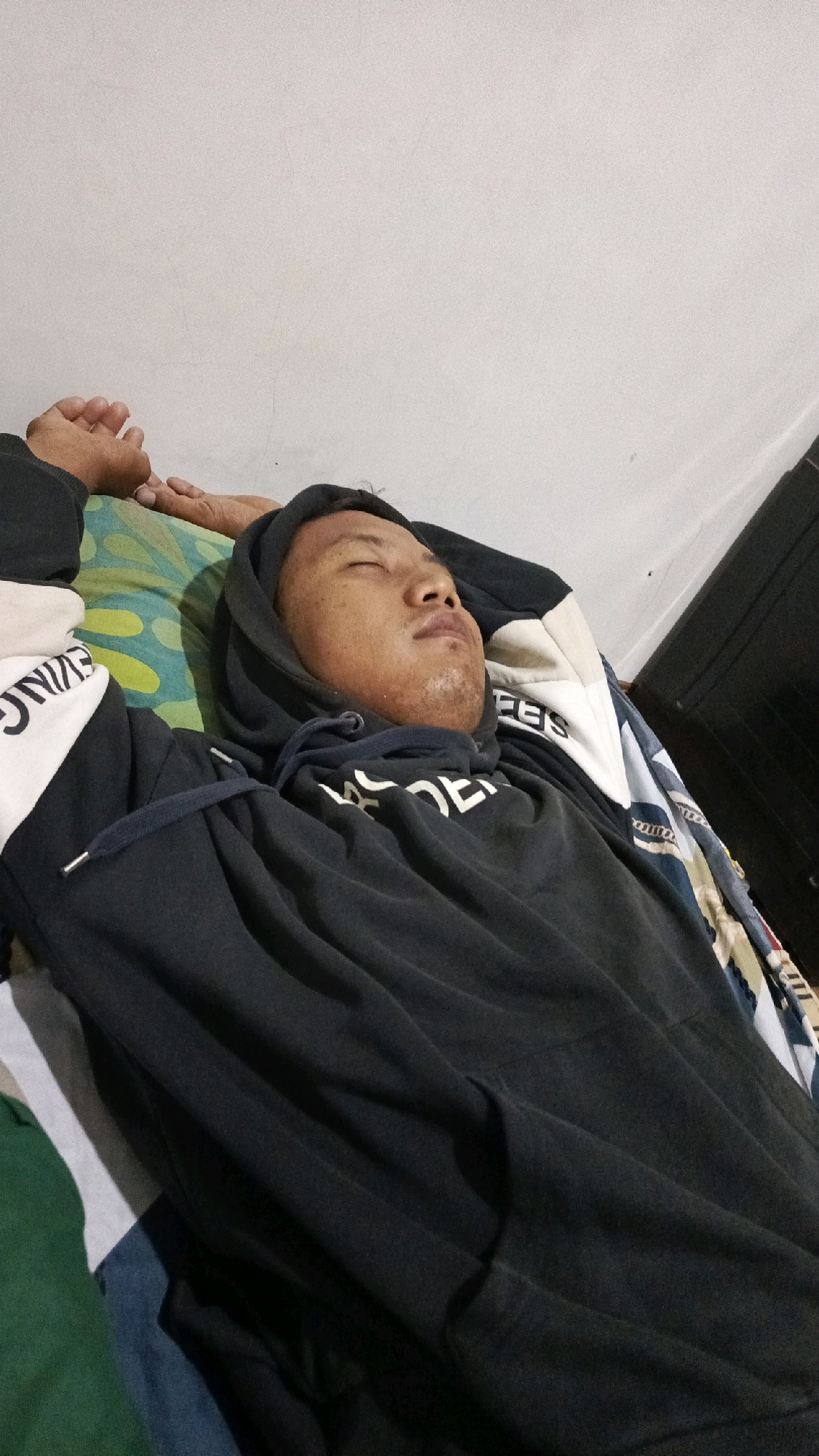 A person is sleeping on a bed in a room. The person is wearing a black hoodie and is lying on their back with their arms resting above their head. The bed has a patterned mattress and various blankets or sheets are visible. The walls of the room are a light color. The image appears to be taken in a residential setting in Pekauman Kulon, Indonesia.