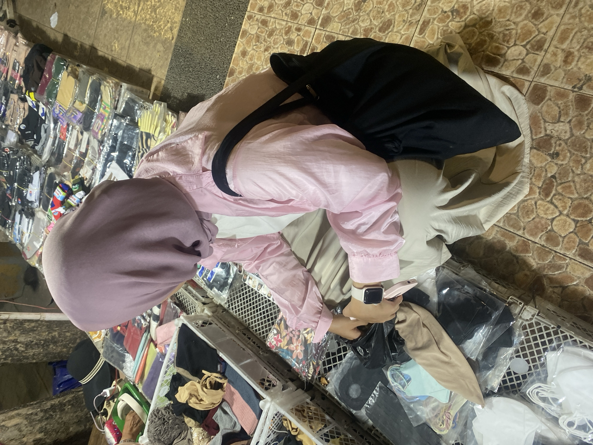 A person wearing a pink hijab and a light pink jacket is kneeling at an outdoor market stall in Banjarmasin, Indonesia. The stall displays various clothing items, including socks and masks, neatly packaged in plastic. The ground is paved with brown tiles. The person is holding a black plastic bag and examining items within a white plastic crate. A black tote bag is slung over their shoulder.