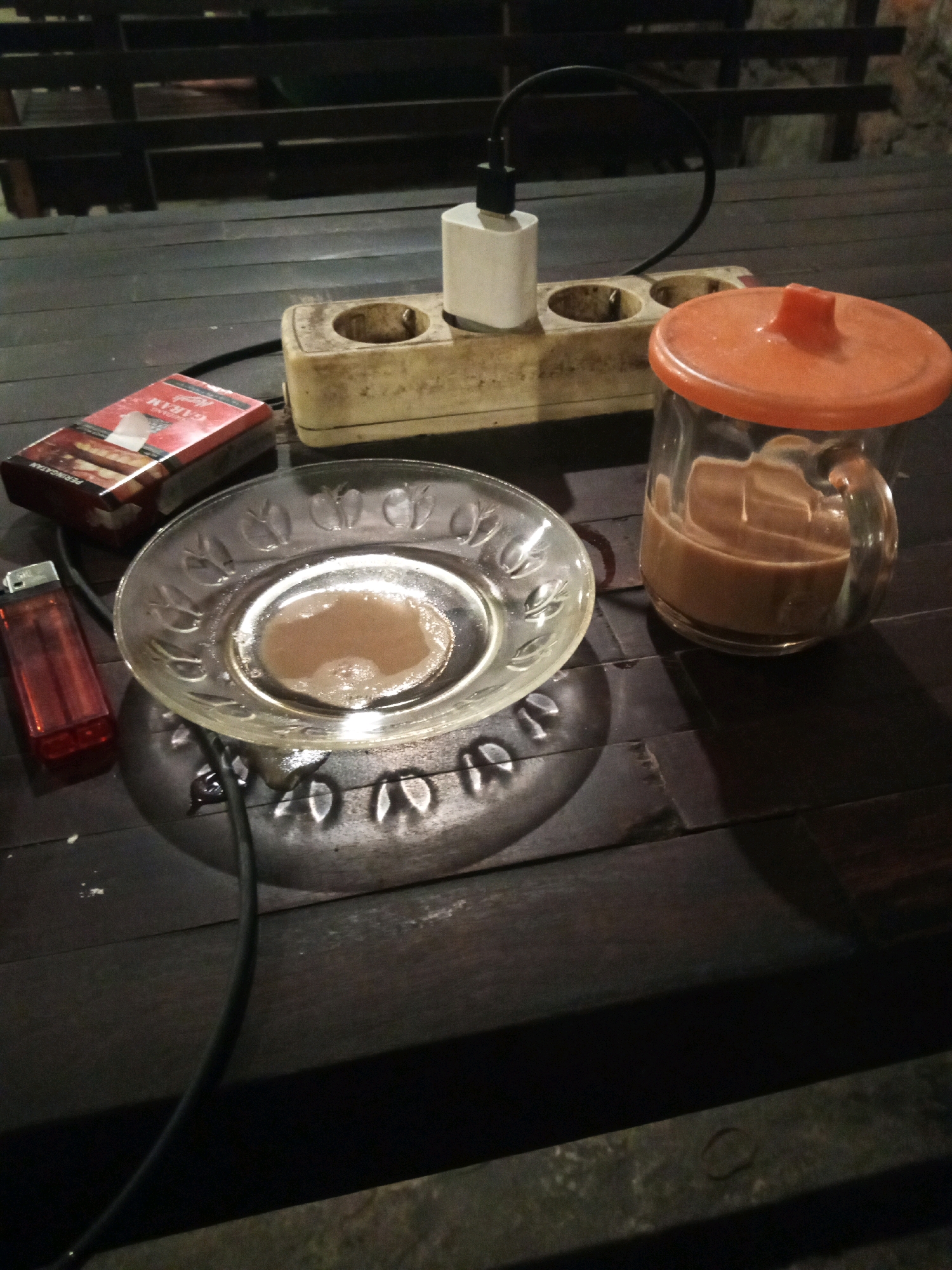The image depicts a dark wooden table with several objects. On the left, there is a red cigarette lighter and a pack of cigarettes with the brand name "GUDANG GARAM" visible. To the right of the cigarettes, there is a clear glass plate with some brown liquid residue. Further to the right, a glass pitcher filled with a brown liquid, likely a beverage, is topped with an orange lid. Behind these items, a white power adapter is plugged into an old, beige electrical outlet strip. A black cable extends from the adapter. The table appears to be outdoors, possibly in a shaded area, with what looks like wooden benches or railings in the background. The lighting suggests it is nighttime or a dimly lit environment. No people are visible in the frame. This scene is located in Tambakagung, Indonesia.