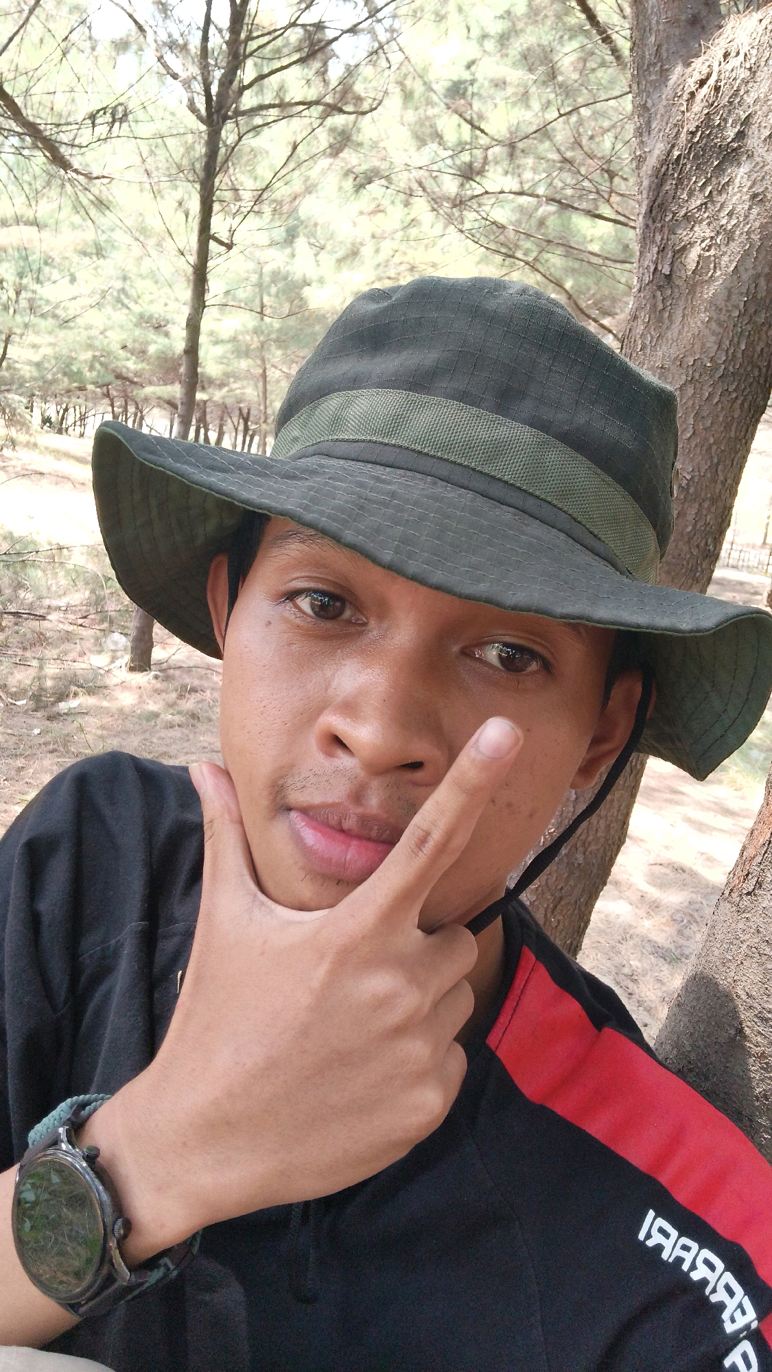 The image is a close-up, outdoor selfie of a young man wearing a dark green bucket hat and a black shirt with a red stripe. He is leaning against a tree with his right hand on his chin and his left hand in front of his face, with his index finger extended. The background consists of trees and dry vegetation, suggesting a natural, possibly rural or park-like setting. The lighting is natural daylight. The man appears to be in good health. The image quality is adequate for identification. There is no indication of the specific location within Pabeanilir, Indonesia, but the environment is consistent with a tropical or subtropical climate.