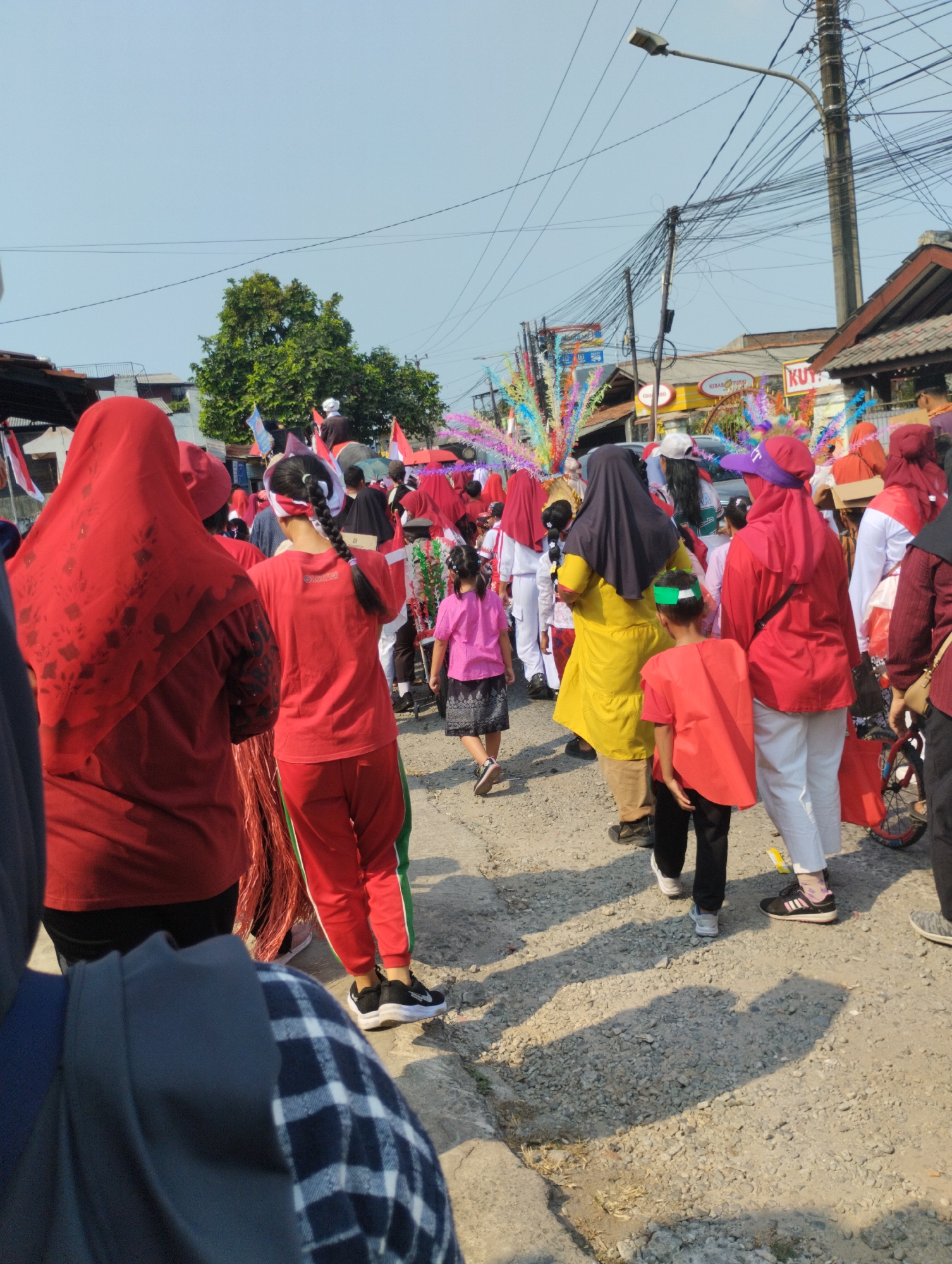 The image captures a street scene in Bekasi, Indonesia, likely during a cultural event or parade. A group of people, predominantly women and children, are gathered on a gravelly road. Many are wearing red attire, including headscarves and outfits, with some donning traditional Indonesian colors. Decorative elements, such as colorful streamers and flags, are visible, suggesting a festive atmosphere. In the background, buildings and utility poles line the street, indicating an urban or semi-urban setting. The bright daylight suggests the photograph was taken during the day.