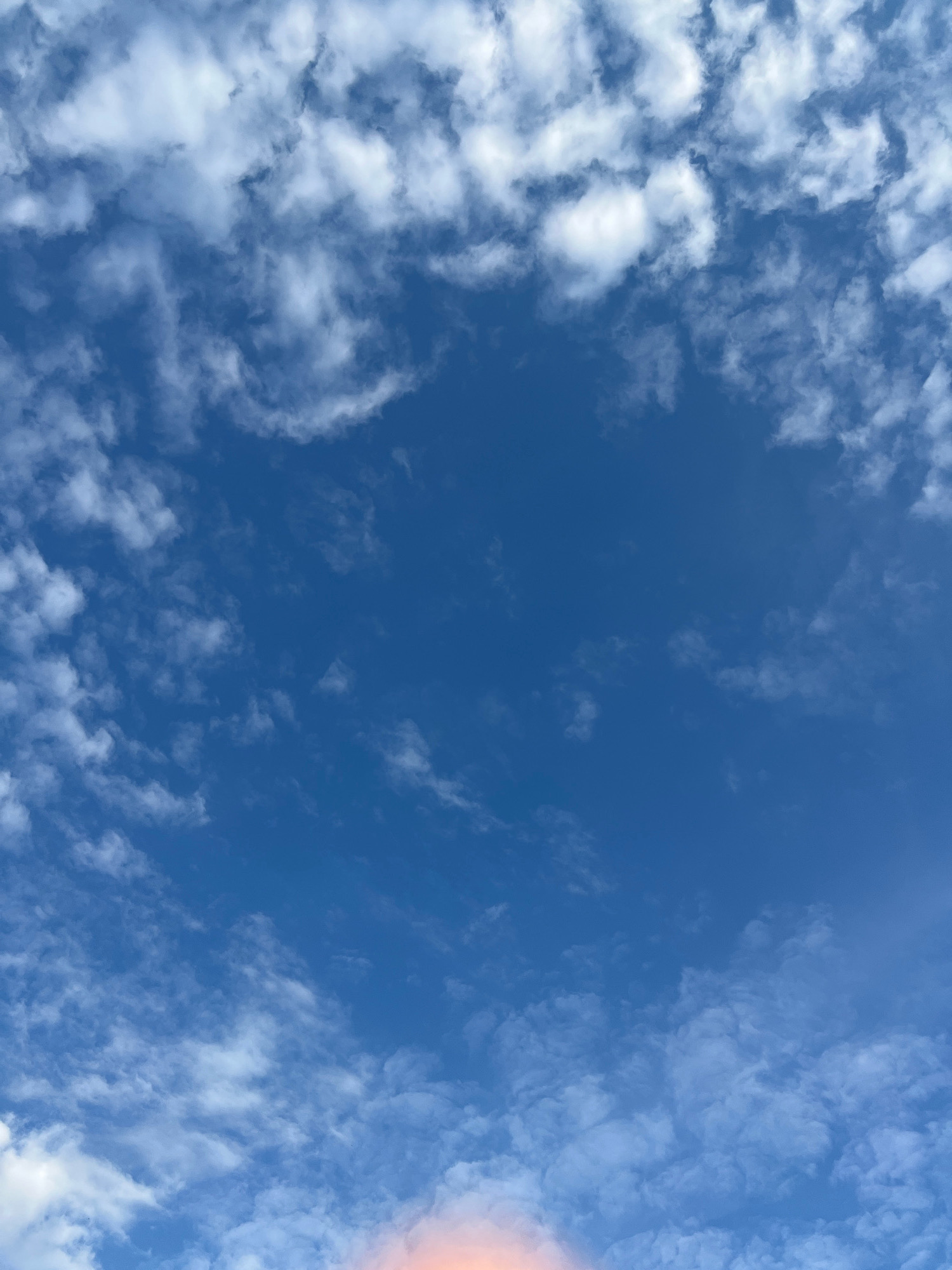 The image depicts a sky with scattered white clouds against a vibrant blue background. The clouds are wispy and varied in density, with some appearing thicker and more defined while others are lighter and more diffused. There's a faint hint of orange or pink light at the very bottom of the frame, suggesting the possibility of a sunrise or sunset. The scene is entirely natural, focusing on the sky and clouds. No specific location within Krasnoyarsk, Russia, can be identified from the image.