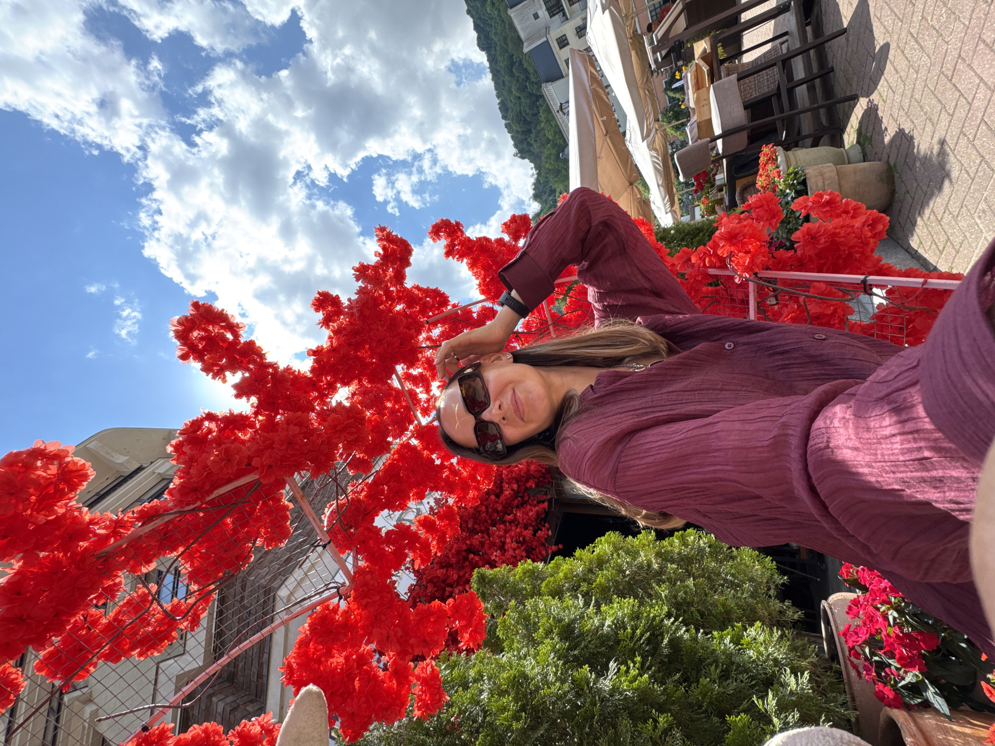 The image depicts a woman posing outdoors in front of a building adorned with vibrant red artificial floral decorations. The woman is wearing sunglasses and a maroon-colored shirt with a crinkled texture. She is touching her hair, looking at the camera with a slight smile. In the background, there are green evergreen trees, potted red flowers, and outdoor seating with umbrellas, suggesting a cafe or restaurant setting. The sky is blue with scattered clouds. The building in the background appears to be of modern architecture. This scene is located in the city of Estosadok, Russia.