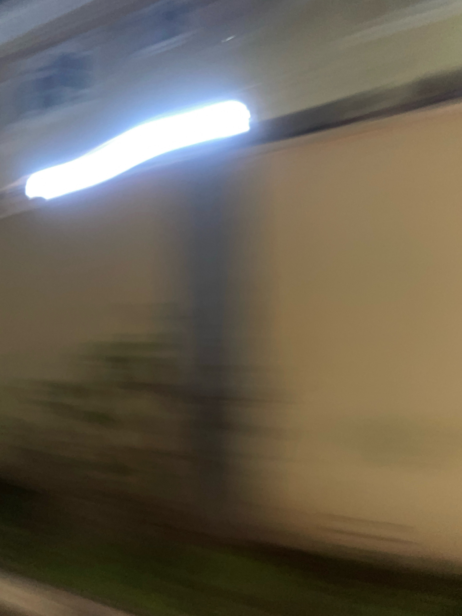 The image depicts a blurred, horizontal view of what appears to be a tunnel or an underpass. A bright, elongated light source, possibly fluorescent lighting, dominates the upper left portion of the frame, casting a strong glare. Below the light, a pale, textured wall stretches across the image, with a darker, vertical shadow or architectural element breaking its uniformity in the center. The lower portion of the image shows a darker, indistinct area that could be the ground or a lower level, with some suggestion of green, perhaps vegetation or painted surfaces. The overall impression is one of movement, possibly captured from a vehicle, given the motion blur. The scene suggests an urban infrastructure setting, consistent with locations in Abuja, Nigeria, such as a pedestrian underpass or a highway tunnel. There are no people or discernible subjects in the image.