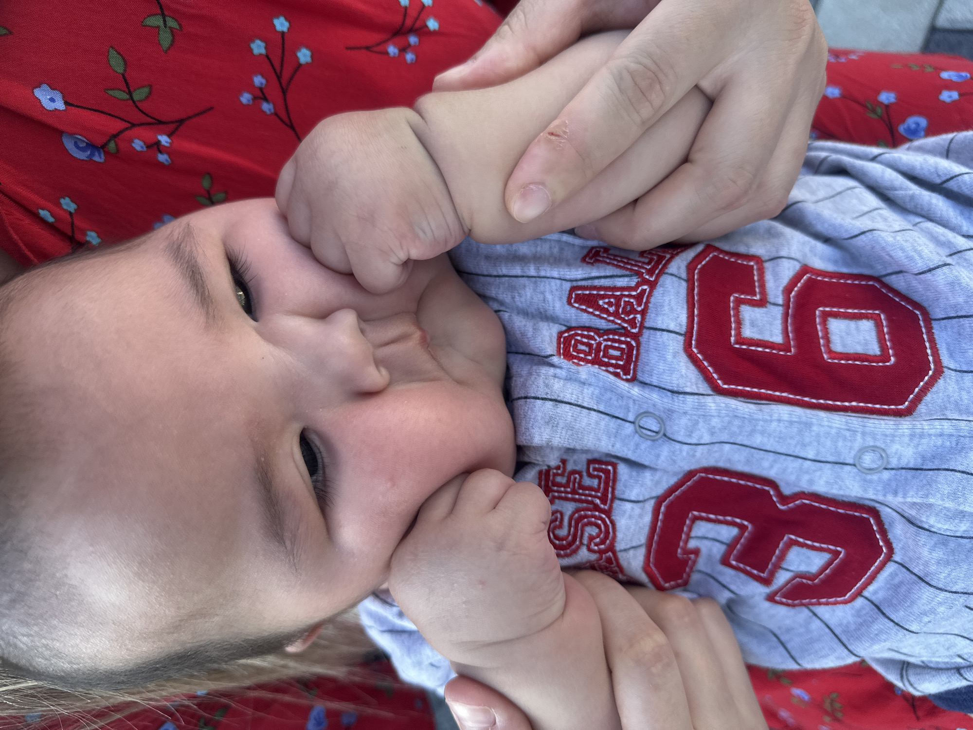 The image shows a close-up, low-angle shot of a baby being held. The baby is wearing a grey and white striped onesie with red lettering and numbers. The onesie has a "6" prominently displayed. The baby has their hands near their face, with one hand appearing to be in their mouth. Adult hands are also visible, gently holding the baby's hands and body. The baby's skin is fair, and their eyes are open and looking towards the left of the frame. The background shows a portion of a red fabric with a floral pattern. The lighting suggests it is daytime, likely outdoors or near a window, as indicated by the natural light. The overall mood conveyed is one of tenderness and care. There is no explicit information about the specific time of day, weather, or precise location within Manas, Kyrgyzstan, beyond the general context provided. The visible text on the onesie includes the number "6" and parts of other letters or words, such as "ASE" and "BALL".