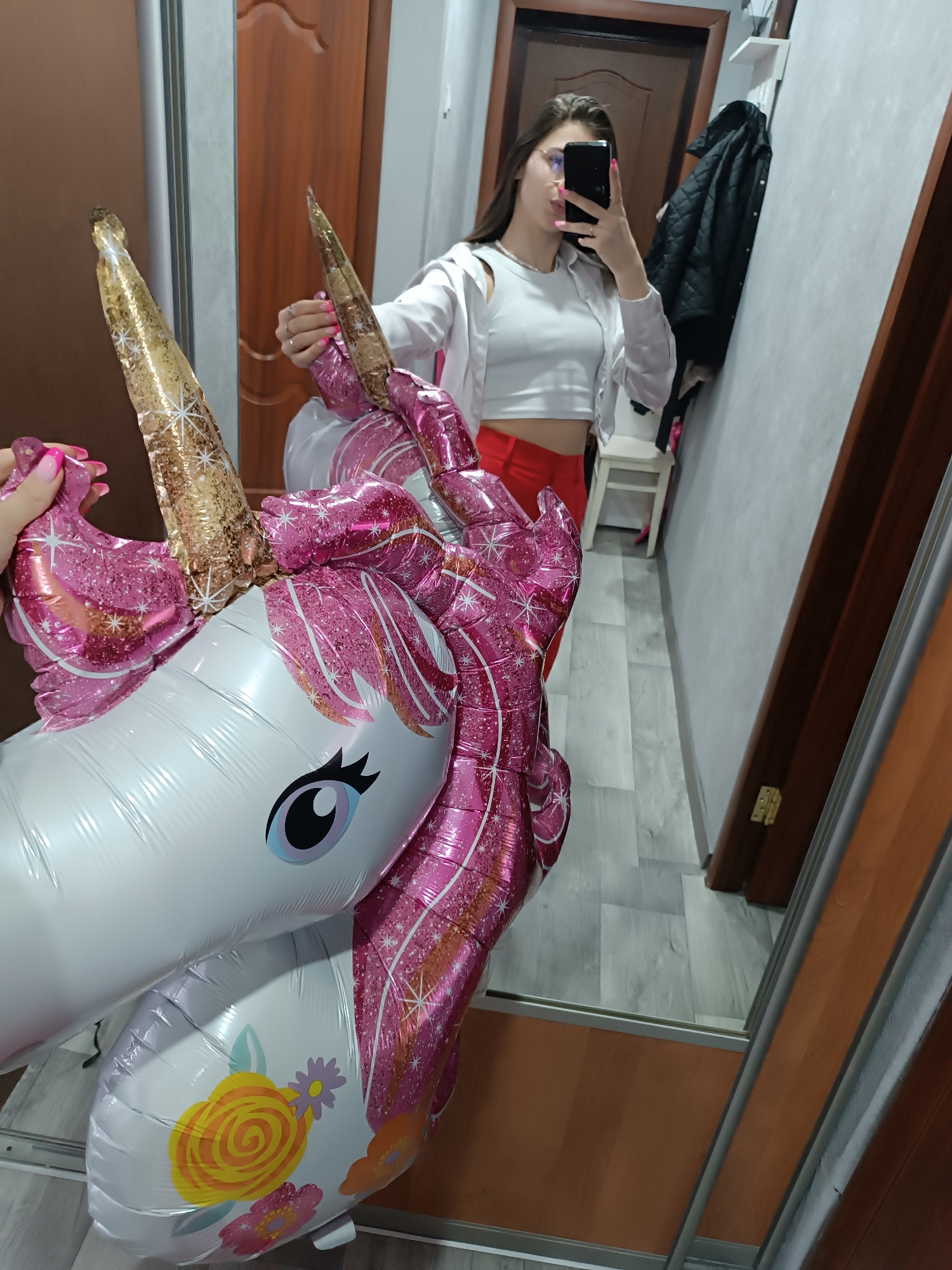 An indoor scene in Unknown, Unknown, shows a female individual taking a mirror selfie. The subject wears glasses, a white crop top, an unbuttoned white shirt, and red pants. Her face is obscured by a black smartphone held in her left hand, which displays pink manicured nails and a silver ring. Her right hand, also with pink manicured nails, supports a large, inflated foil unicorn balloon. The balloon is predominantly white with a glittery pink mane and tail, a glittery gold horn, blue and black eyes, and multicolor floral decals on its lower body. The reflection reveals a room with light grey plank flooring. A dark brown wooden door is visible on the left and right, with a brass hinge on the right door frame. A light grey wall, a white wall-mounted shelf, a black quilted jacket on a hook, and a small white stool are also present in the background.