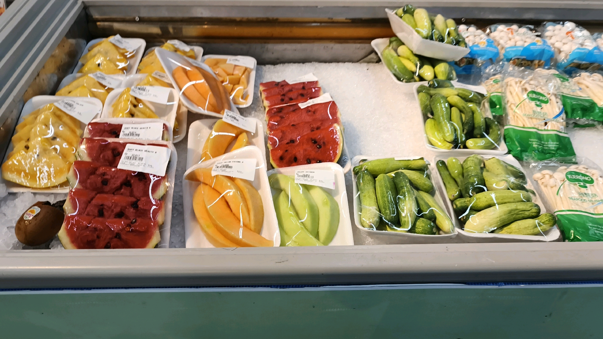 The image depicts a refrigerated display case in a market, likely in Baturaja, Indonesia, filled with various fruits and vegetables pre-packaged in plastic. The display case is divided into sections on ice. In the foreground, on the left, are slices of yellow fruit, possibly pineapple or mango, and several slices of watermelon. To the right of the watermelon are slices of a light orange fruit, likely cantaloupe, and slices of green melon. Further to the right are rows of cucumbers, and in the background are packages of mushrooms. The overall environment appears to be a commercial setting, possibly a grocery store or wet market, focused on displaying fresh produce.