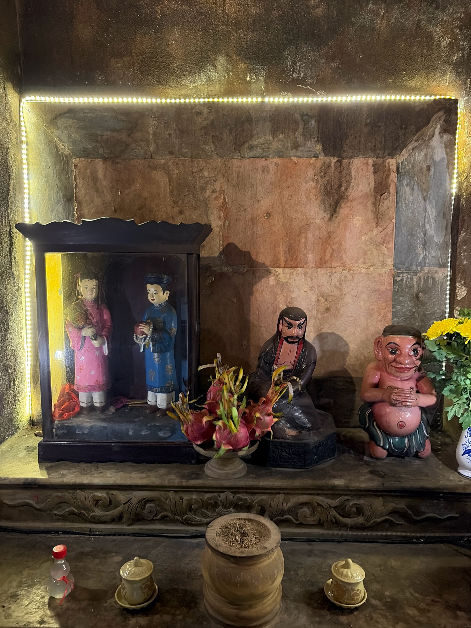 The image shows an altar with several figurines. In the center is a display case containing two figurines dressed in traditional Vietnamese clothing. To the right of the display case, there are two larger figurines. One is seated and appears to be a warrior, while the other is plump and smiling. In front of the figurines are some offerings, including flowers and small ceramic cups. The altar is lit by string lights around the background. The overall setting appears to be within a temple or shrine in Da Nang, Vietnam, given the context of the request.
