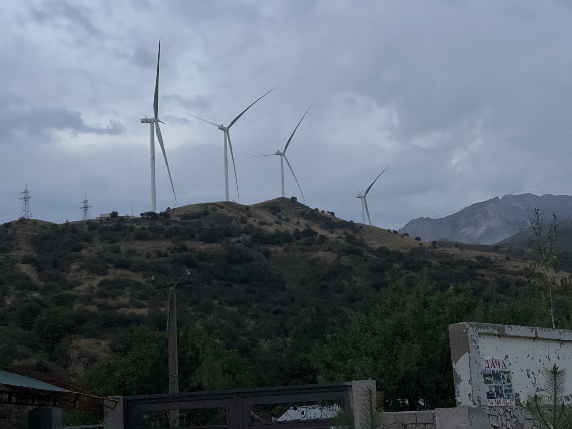 In Yusuphana, Uzbekistan, on a cloudy day, several wind turbines stand on a grassy hill. The turbines are white with slender blades that point upwards. The hill is covered in dry grass and scattered green bushes. In the foreground, there are trees and a low wall. To the right, a white building with peeling paint has a poster on it with Cyrillic text. In the background, mountains are visible. Several power lines and poles are also present on the hill.