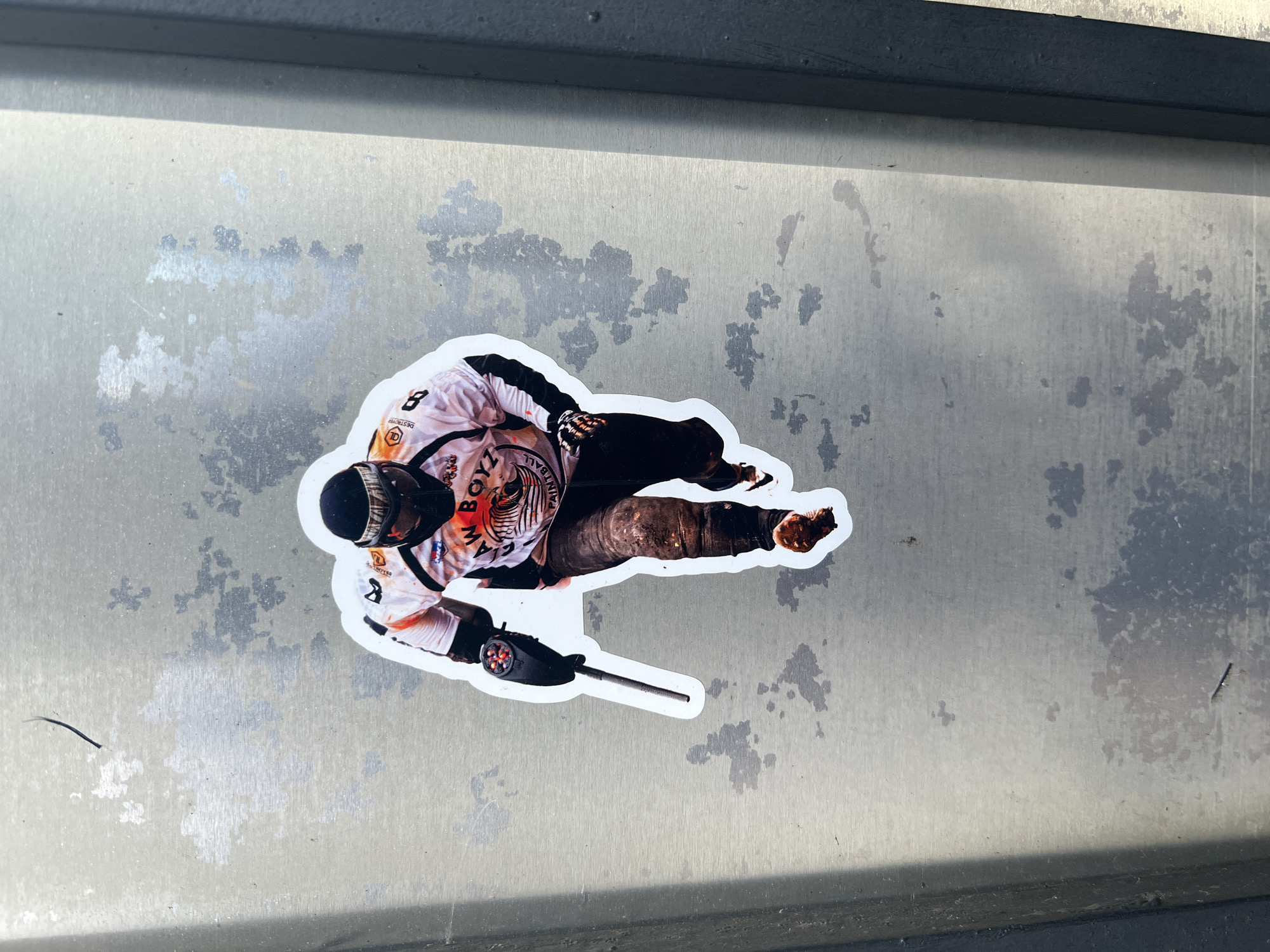 A sticker depicting a paintball player is adhered to a metal surface in the City of Newburgh, United States of America. The sticker shows a figure in paintball gear, including a mask, jersey (with "RAW BOYZ PAINTBALL" printed on it), pants, and a paintball marker. The player is positioned low to the ground, as if in action. The metal surface has a mottled texture with areas of discoloration and wear.