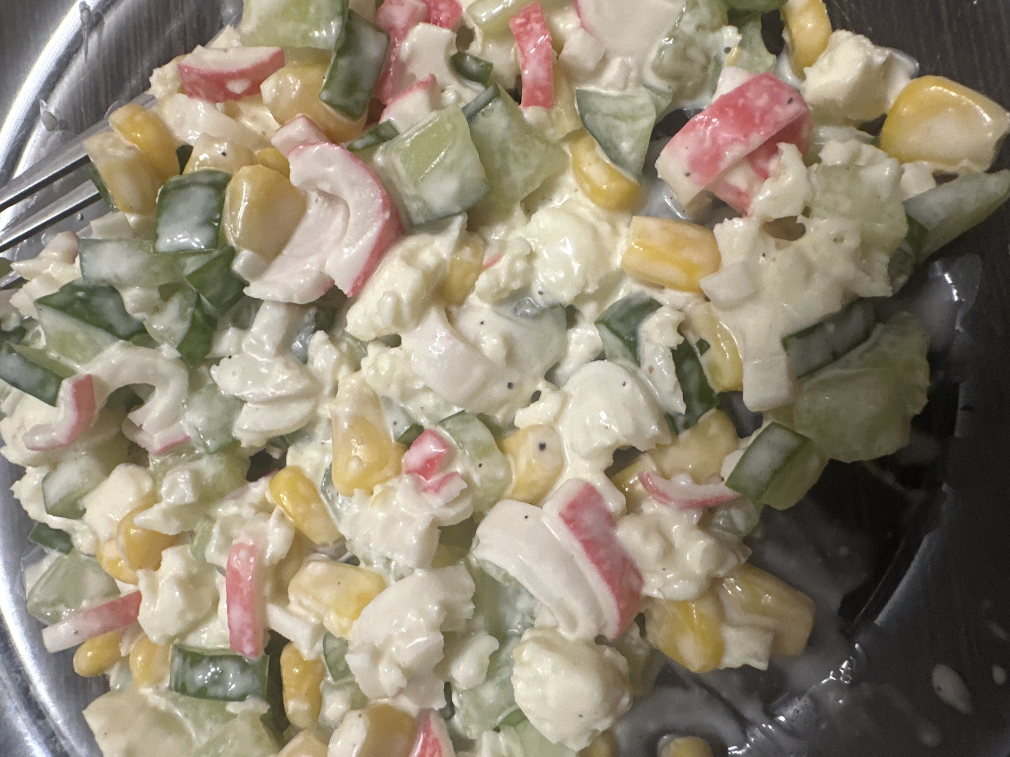 The image shows a close-up, overhead view of a salad in a dark bowl. The salad appears to be creamy and consists of chopped ingredients including corn, cucumber, and imitation crab meat. A fork is partially visible in the upper right corner. The salad is likely prepared at home. The scene is not specific to Novoaltaysk, Russia, and no information about the location can be determined from the image.