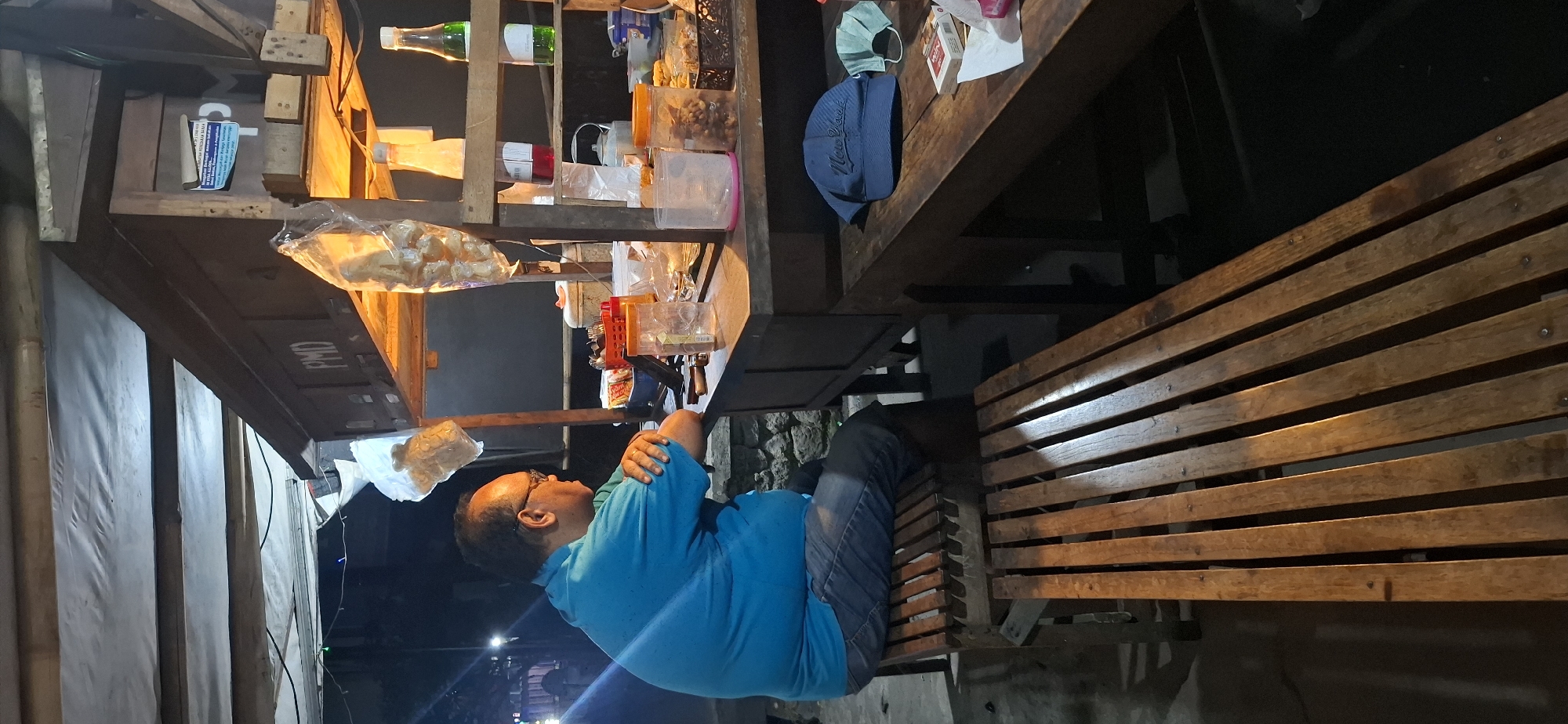 A middle-aged man with glasses is sitting on a wooden bench in front of a food stall in Colomadu, Indonesia. The stall is lit with warm lighting, and there are various food items and condiments displayed on shelves. The man is wearing a blue t-shirt and jeans, and he appears to be looking at something on the counter. The bench he is sitting on is made of weathered wood slats. In the foreground, another wooden bench is visible. The overall scene suggests a casual, nighttime setting at a local eatery.