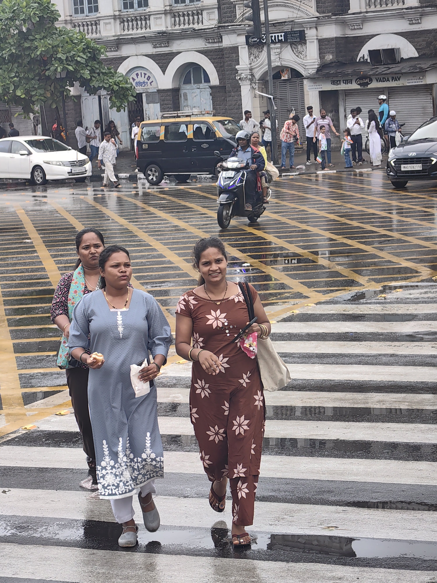 The image depicts a busy street scene in Mumbai, India, likely near the iconic CSMT (Chhatrapati Shivaji Maharaj Terminus) station, given the architectural style of the buildings and the general ambiance. The foreground shows a group of three women crossing a wet pedestrian zebra crossing, with yellow lines demarcated on the asphalt. The woman in the center is wearing a light blue kurta with white floral embroidery and holding a small pastry and a white napkin. The woman to her left wears a dark patterned blouse with a colorful scarf. The woman to her right is wearing a brown kurta with a white floral pattern and carrying a light-colored bag over her shoulder. They are walking from left to right across the frame.
In the background, several vehicles are visible, including a white car, a yellow and black taxi, and a scooter with two riders. Pedestrians are also scattered throughout the scene, some standing and others walking. The buildings in the background are of colonial architecture, characterized by arched windows and ornate detailing. The street appears wet, indicating recent rainfall, which is common in Mumbai, especially during monsoon season. The overall atmosphere is one of urban activity, with people and vehicles navigating the city streets.