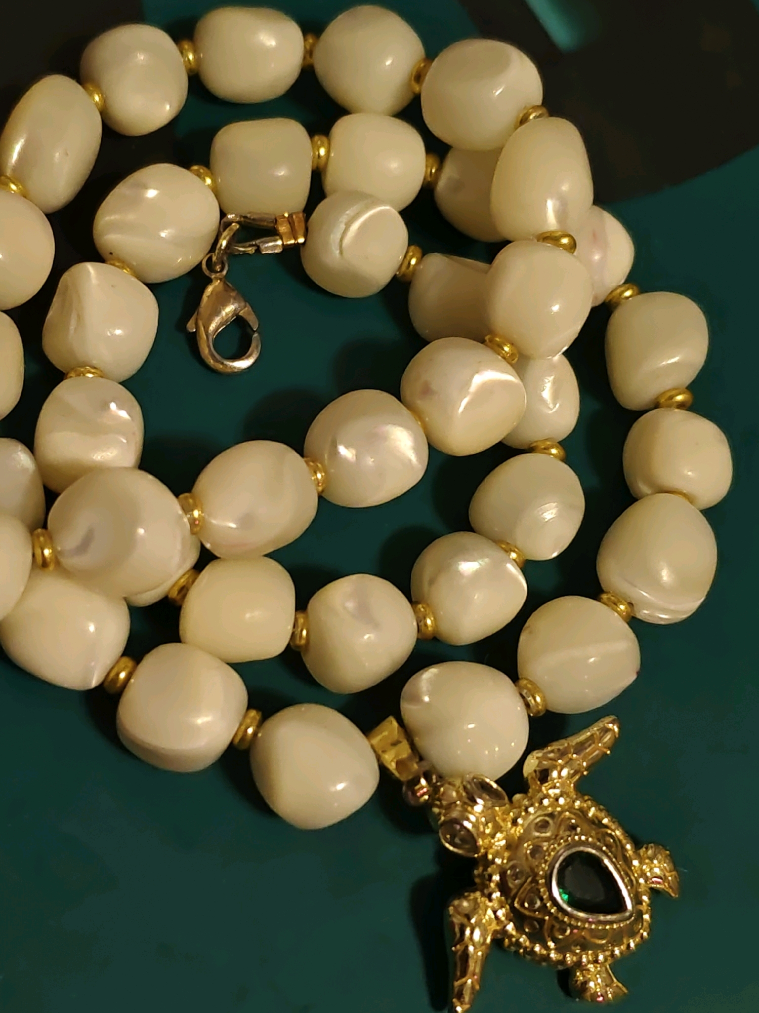 A close-up, overhead shot shows a creamy white beaded necklace with gold accents and a gold turtle-shaped pendant with a dark green stone. The necklace is laid out on a dark teal surface. The background is out of focus, but appears to be the same teal color. The image was taken in Rostov-on-Don, Russia.