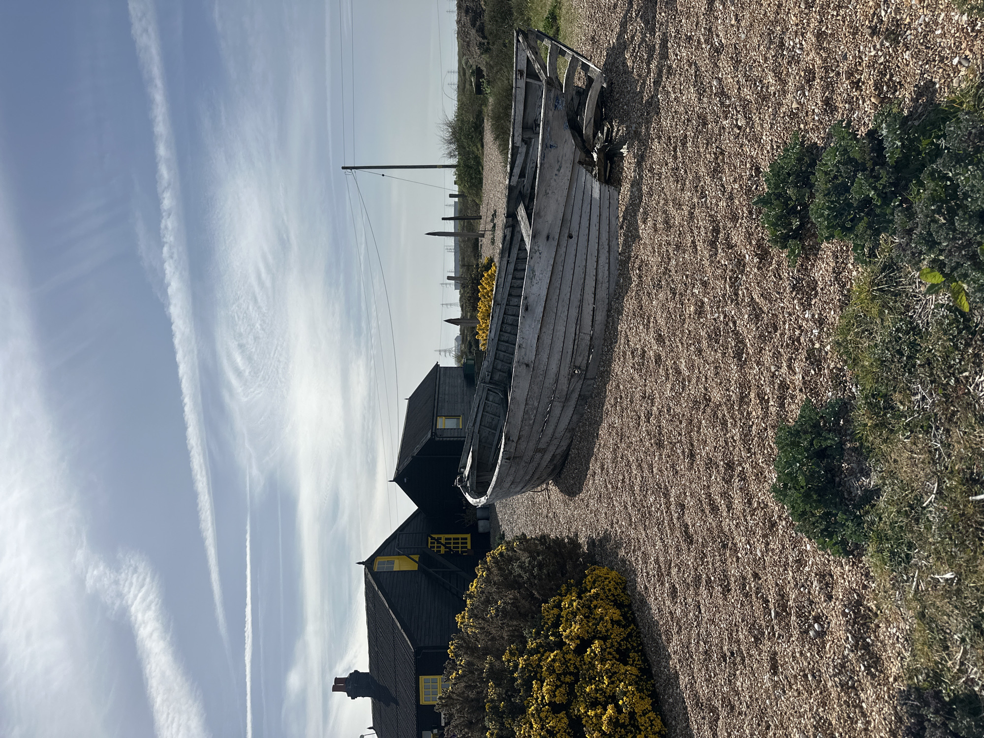 The image shows a scene at Dungeness, Folkestone and Hythe District, United Kingdom. An old, weathered wooden boat rests on a shingle beach, next to an area of vegetation, including patches of green plants and a yellow flowering bush. Behind the boat is a black wooden building with yellow-framed windows. Power lines stretch across the sky, which is light blue with streaks of clouds. In the distance, more buildings and coastal vegetation can be seen. The ground is covered with brown shingle.
