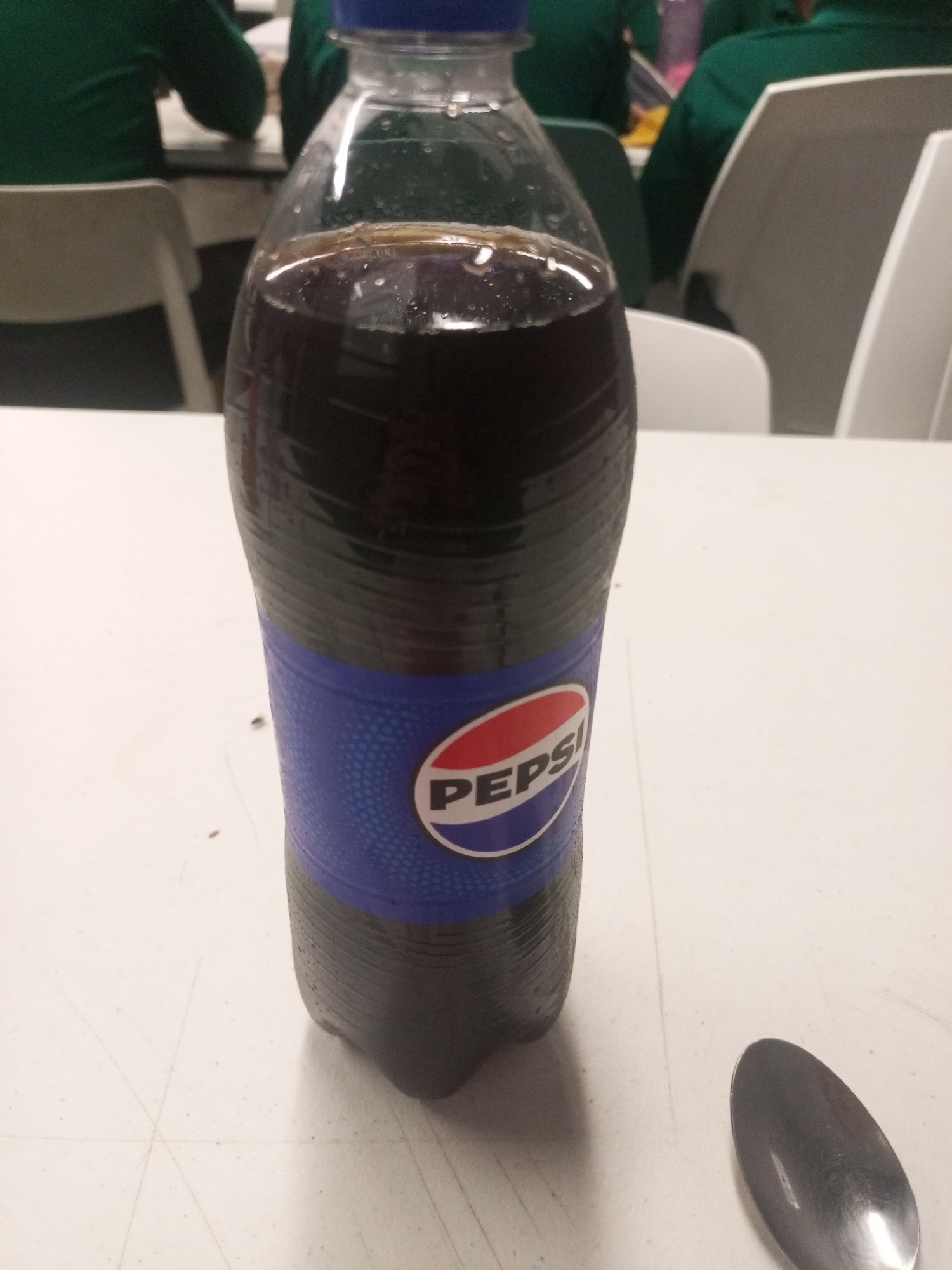A partially full bottle of Pepsi stands on a white table in an indoor setting. The bottle has a blue cap and a blue label with the Pepsi logo. A metal spoon rests on the table to the right of the bottle. In the background, several people wearing green shirts are seated at tables. The location is unspecified, and there is no evidence to suggest the image was taken in Juan Griego, Venezuela.