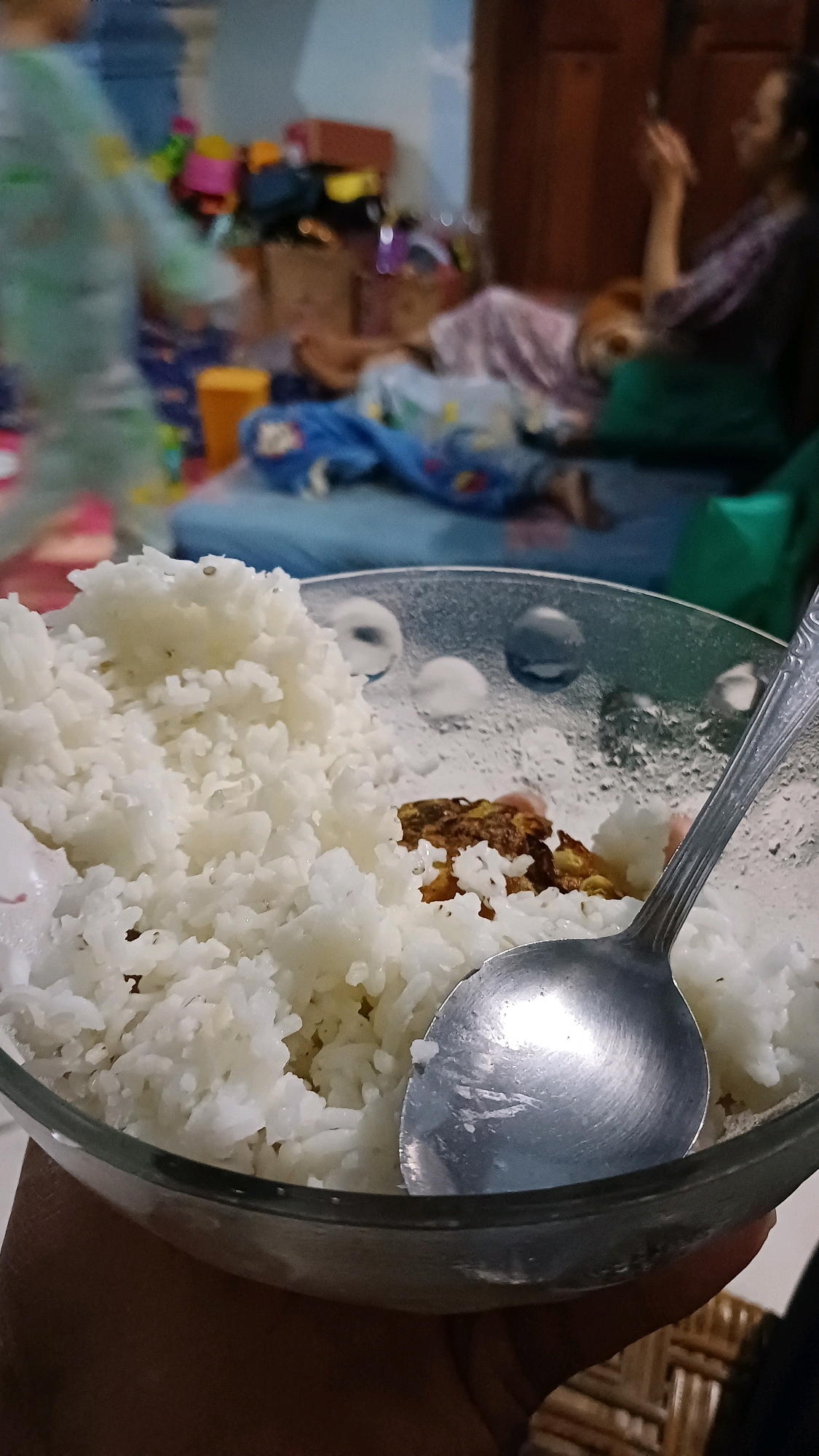 A close-up shot captures a bowl of white rice overflowing with a serving spoon resting on top. The bowl is held by a hand, and the background is blurred, showing a room with people and furniture, suggesting a home environment. The overall impression is that of a simple meal being prepared or consumed. The image is set in the city of Wonokerso, Indonesia.