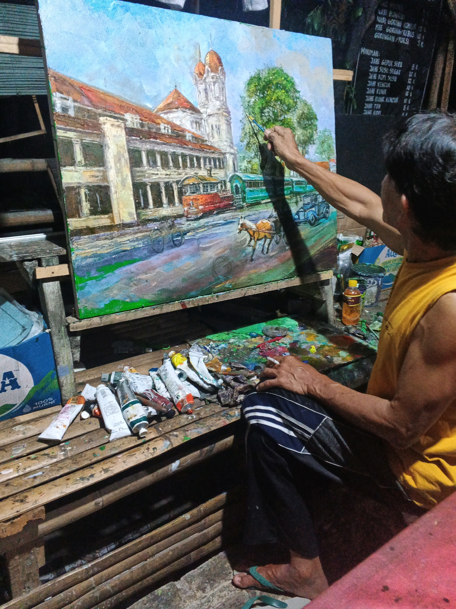 The image shows a man painting a picture of a historical building with a light blue sky and green trees. The man is wearing a yellow sleeveless shirt and dark pants with white stripes. He is holding a paintbrush in his right hand and a cigarette in his left hand. The painting is on an easel in front of him. A palette with various colors is on a wooden surface to his right. Several tubes of paint are scattered on a bamboo surface to his left. In the background, there is a menu board with Indonesian text. The scene appears to be an outdoor art studio, possibly in Ungaran, Indonesia, given the menu board text.