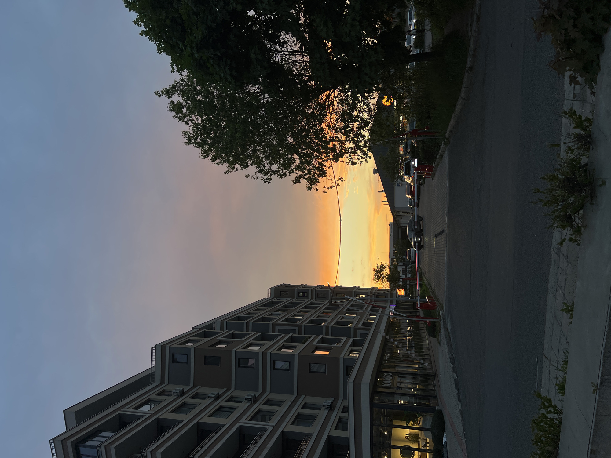 The image depicts a cityscape during sunset. In the foreground is a multi-story building with a modern architectural design, featuring recessed balconies and a mix of dark and light grey hues. The building appears to have commercial spaces at street level with large glass windows. Adjacent to the building is a paved road with parked cars on one side. Above the building, the sky transitions from a light blue to a warm orange and yellow glow, indicating the setting sun. A tree with full foliage is visible on the upper right side of the frame, silhouetted against the bright sky. The building is located in the city of Burgas, Bulgaria.