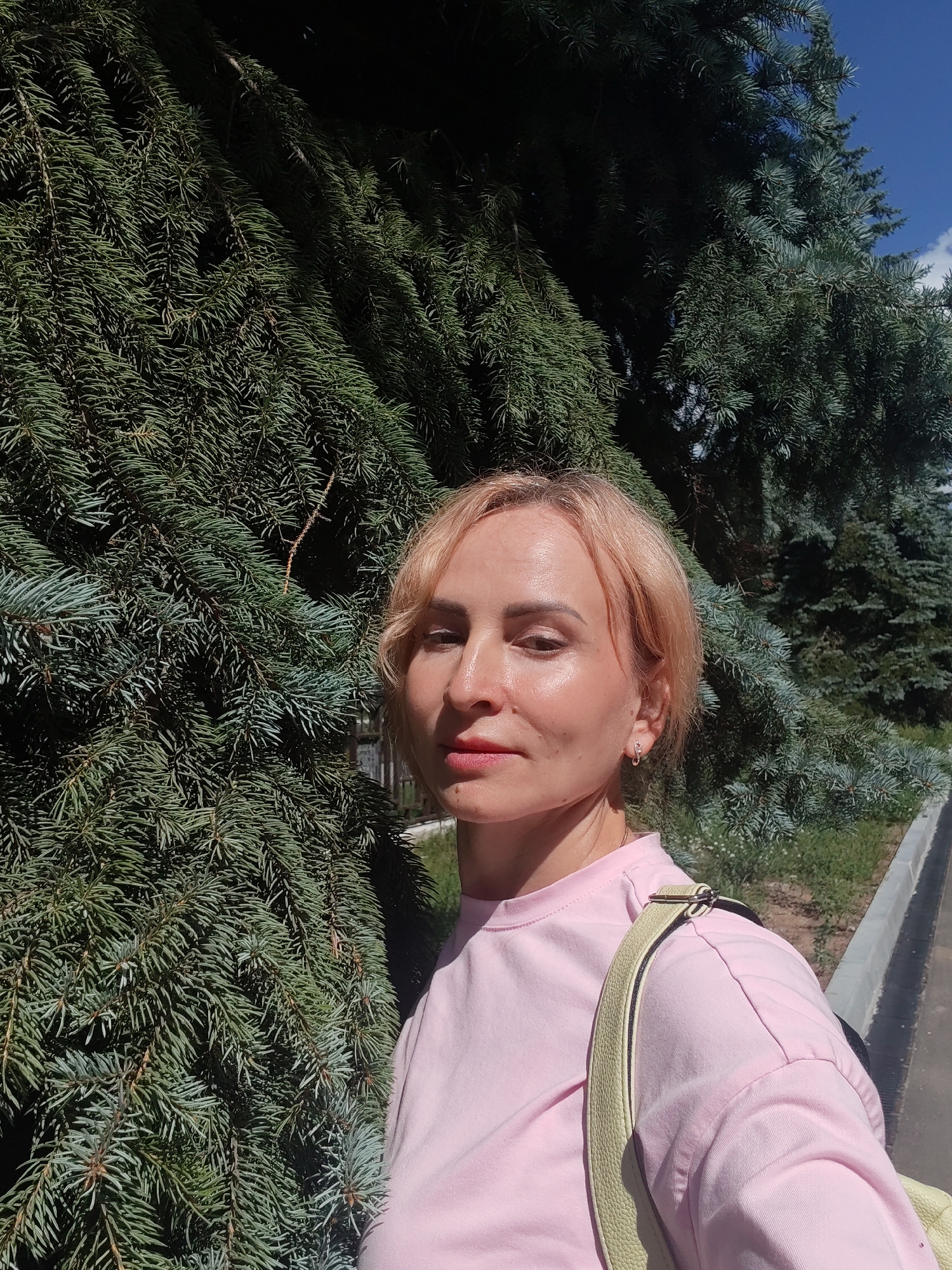 A woman with short blonde hair and fair skin is standing outside in front of a large evergreen tree. She is wearing a pink t-shirt and a light-colored strap from a bag is visible over her shoulder. The background consists of the dense branches of the tree, a concrete curb, grass, and a bright blue sky with some clouds. The lighting suggests it is daytime and sunny. The image appears to be a selfie. The specific location in Chaikovsky, Russia cannot be determined from the visual information alone.