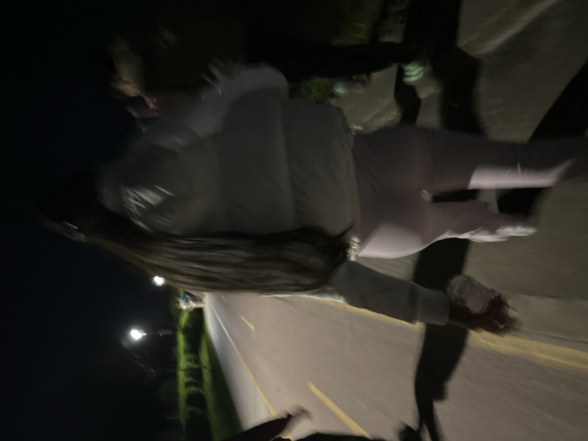 A woman with long, dark hair wearing light-colored clothing, including leggings and a jacket, is walking along a paved path illuminated by streetlights at night. There is greenery visible on one side of the path, and the surrounding area appears dark. There are streetlights illuminating the road. The location appears to be in an urban area, potentially a park or pedestrian zone. The exact location in Plovdiv, Bulgaria, cannot be determined without additional information.