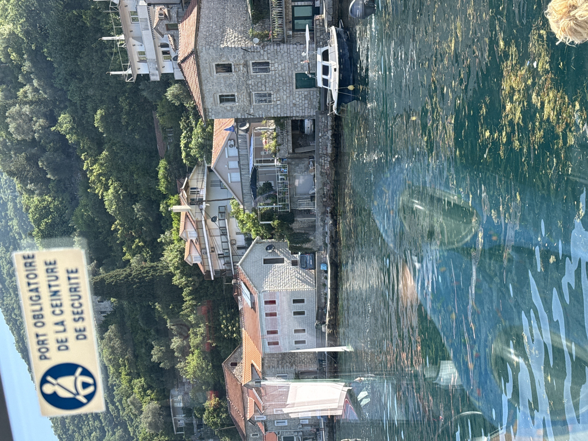 In Lepetane, Montenegro, a sign with a blue circle containing a white silhouette of a person wearing a seatbelt is displayed. The text on the sign reads "PORT OBLIGATOIRE DE LA CEINTURE DE SECURITE," which translates to "Mandatory seatbelt use." The sign is affixed to a surface, likely a window or vehicle interior, and is set against a backdrop of lush green hills and waterfront buildings. The water in the foreground is a deep blue, reflecting the surrounding scenery and the sky. Several boats are visible, including a small white and blue motorboat docked near a stone building. The architecture of the buildings suggests a Mediterranean coastal town. The overall scene depicts a scenic view with a prominent safety reminder.