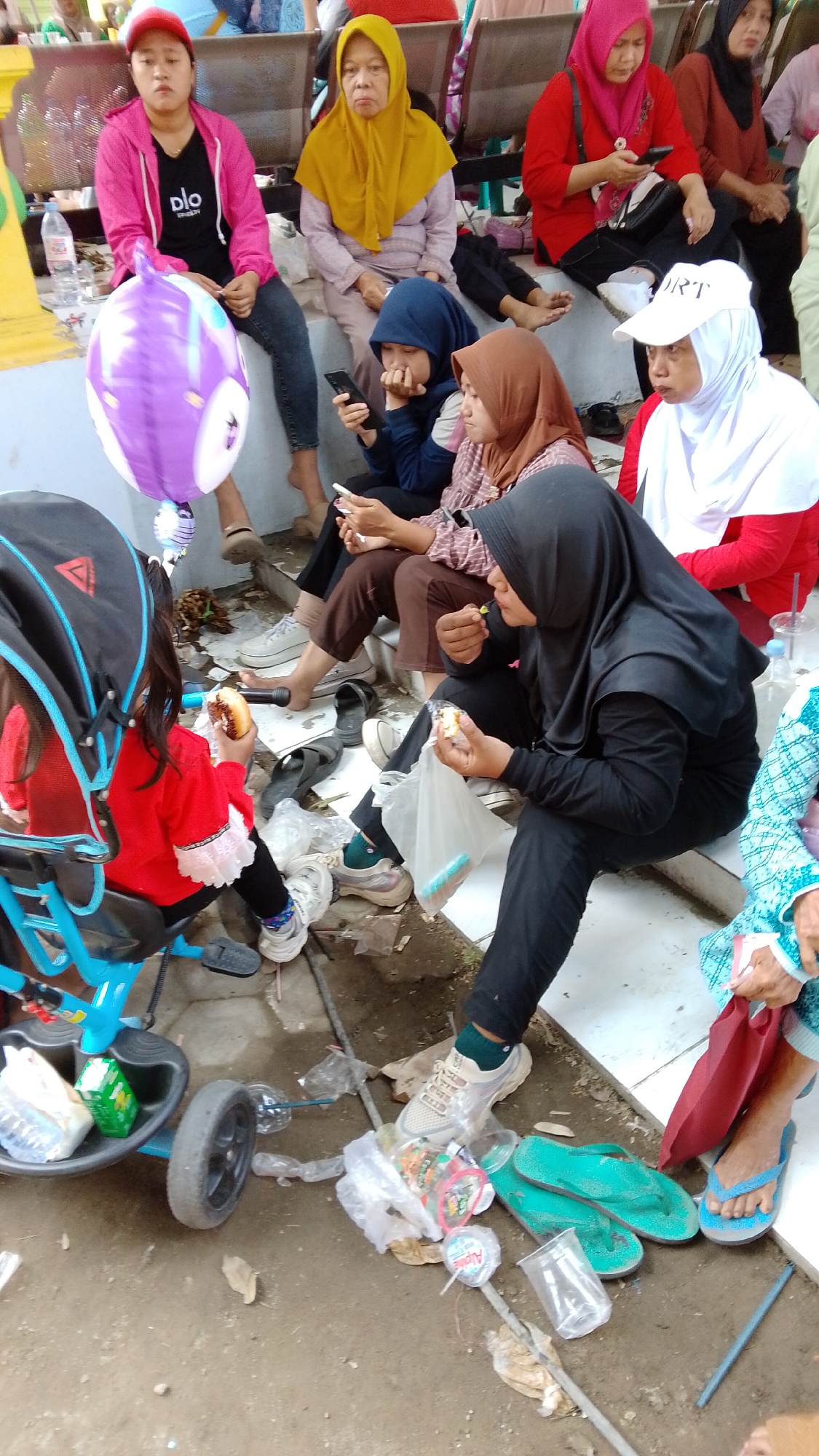 In Mancar, Indonesia, a group of women and a young child are gathered outdoors. One woman, wearing a bright pink hooded jacket and a red cap, sits with a purple balloon. Beside her, an elderly woman in a yellow hijab looks forward. Another woman in a blue hijab is looking at her phone, while a woman in a brown hijab eats from a plastic bag. A woman in a black hijab is also eating. In the foreground, a child in a red top sits in a blue stroller, holding a donut. The ground is littered with plastic bags, cups, and discarded food wrappers. Two pairs of flip-flops, one blue and one dark-colored, are visible on the ground. The scene appears to be set on a paved area with some steps leading up to where the individuals are seated.
