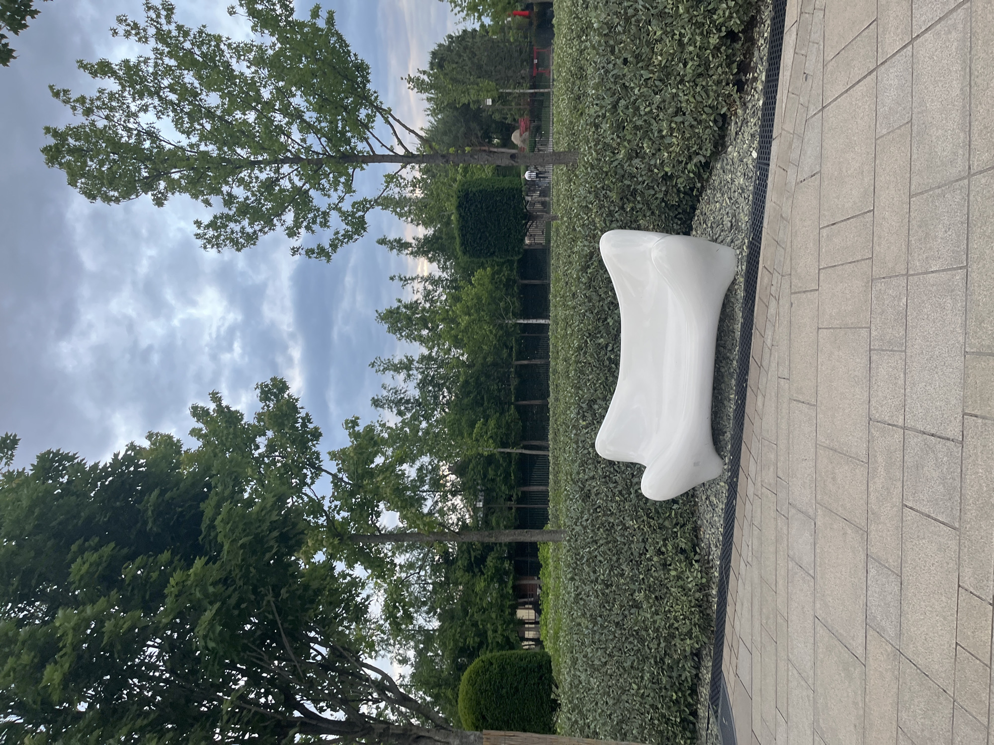 The image shows a white, abstractly shaped outdoor bench located in a park or public space in Krasnodar, Russia. The bench is situated on a paved walkway made of rectangular stone tiles. To the left of the walkway is a landscaped area with a dense hedge of small green bushes. Behind the hedge are several rows of trees with green foliage. In the background, more trees and structures are visible. The sky is partly cloudy.