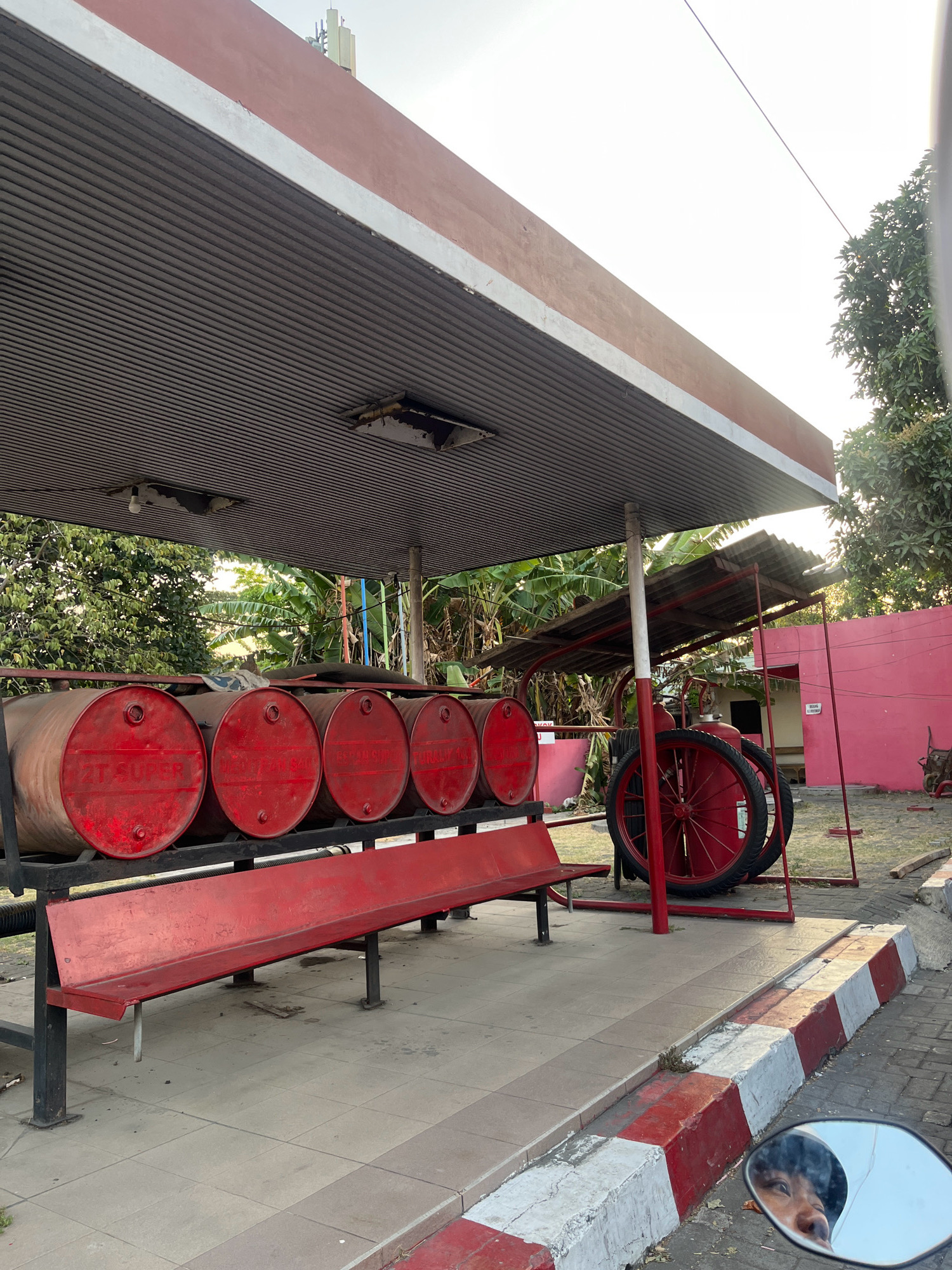 A red metal gas station canopy covers a red metal bench, five red metal barrels, and a red wheeled cart. The barrels are labeled with white text including "2T SUPER". Behind the barrels, a building with pink walls is visible. The ground is tiled and bordered by red and white curbs. A motorcycle mirror in the lower right corner reflects the face of a person. The scene is located in Calukan RT 1 RW 5 Keboansikep, Indonesia.