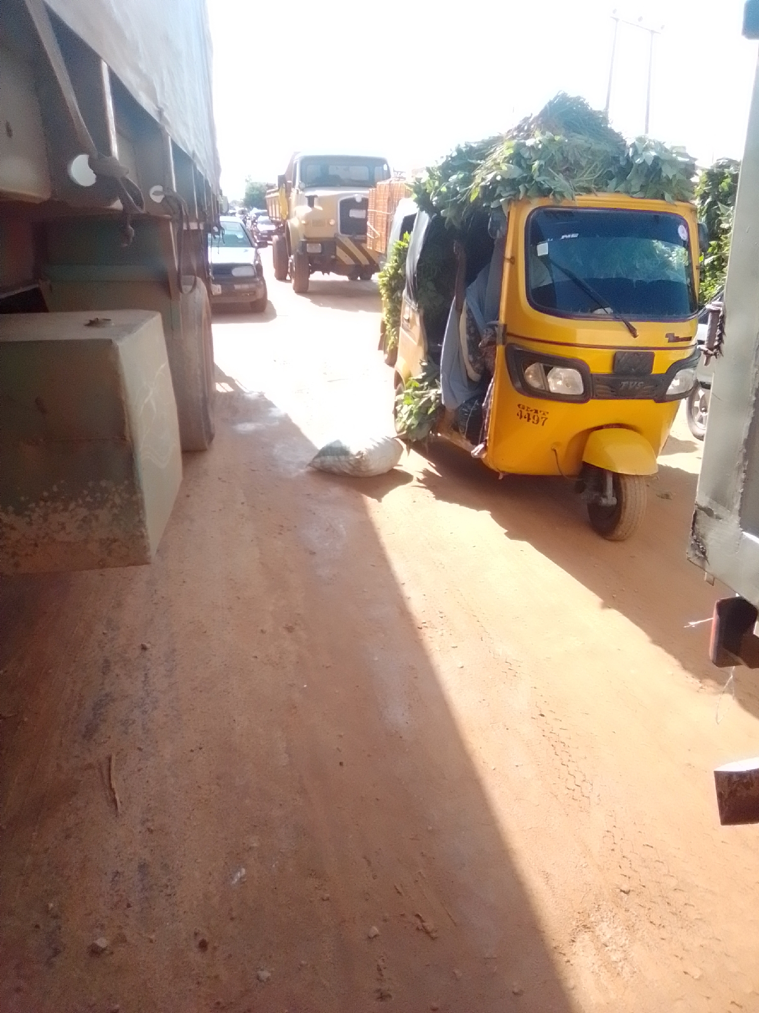 The image depicts a dusty road in Kwadon, Nigeria, with several vehicles present. In the foreground on the left, a large truck's trailer is partially visible, occupying a significant portion of the frame. To the right of the truck, a yellow auto-rickshaw, also known as a "Keke" or "Maruwa," is laden with a large quantity of green foliage, likely produce, piled high on its roof and sides. A person is partially visible inside the auto-rickshaw, driving it. Behind the auto-rickshaw, a yellow truck is visible, and further in the background, other vehicles and a pedestrian can be seen. The road surface is composed of reddish-brown dirt and sand, with tire tracks visible in places. The sky is bright and overcast, suggesting daylight. The overall scene suggests a busy, possibly market or transportation hub, area.