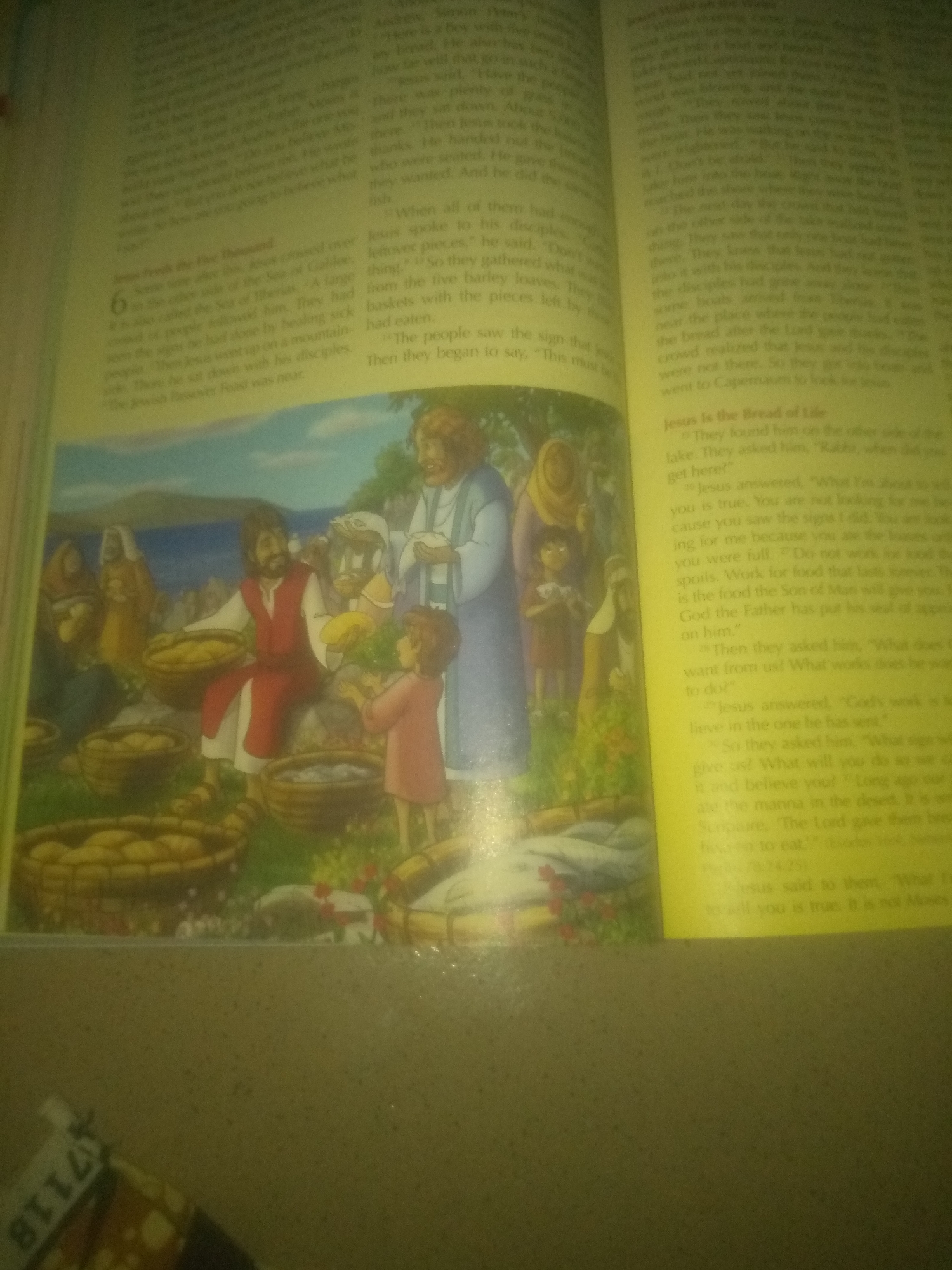The image shows an open book with text and an illustration. The text appears to be from a religious scripture, likely the Bible, with headings and verses. The illustration depicts a scene with several people gathered outdoors, with baskets of bread and fish. One man, likely Jesus, is distributing food, and a child is offering bread. The background shows hills and a body of water. The lower portion of the image shows the corner of another object with the numbers "7118" printed on it. The overall lighting suggests the image was taken indoors, with a yellow tint. The location in Jauro Yinu, Nigeria, is not discernible from the image itself.