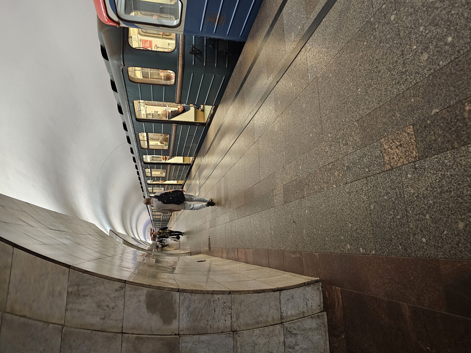 This is a photograph of a metro station platform in Moscow, Russia. The station features a curved ceiling with a white, textured surface. The walls are lined with large, light-colored marble tiles, and the floor is paved with granite tiles in shades of gray and brown. A train with teal-colored carriages and red and blue accents is stopped at the platform, with its doors open. Several people are visible on the platform and inside the train. One man in a light-colored shirt and jeans, carrying a black backpack, is walking away from the camera down the platform. Another person, wearing a dark jacket and carrying a handbag, is walking in the opposite direction. The lighting in the station is artificial, creating shadows on the platform.