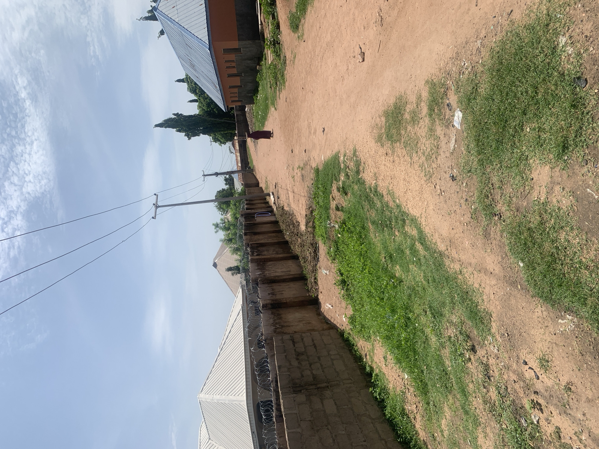A person wearing a dark garment walks away from the camera on a dirt road in Jauro Yinu, Nigeria. The road is flanked by a low, concrete block wall on the left, topped with barbed wire. Beyond the wall are houses with corrugated metal roofs. On the right side of the road, there is a building with an orange facade and a blue metal roof. Trees, including tall, slender evergreens, are visible in the background. Overhead, power lines stretch across a bright sky with scattered white clouds. The ground is a mix of dirt and sparse green grass, with some litter scattered along the road.