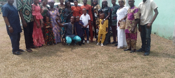 A large group of people stands on a grassy lawn. Many adults wear colorful, patterned clothing. There are also several children present, some standing and others being held or posed with by adults. One man is kneeling and holding up a phone, presumably taking a group photo. The background features a light-colored building or structure.
