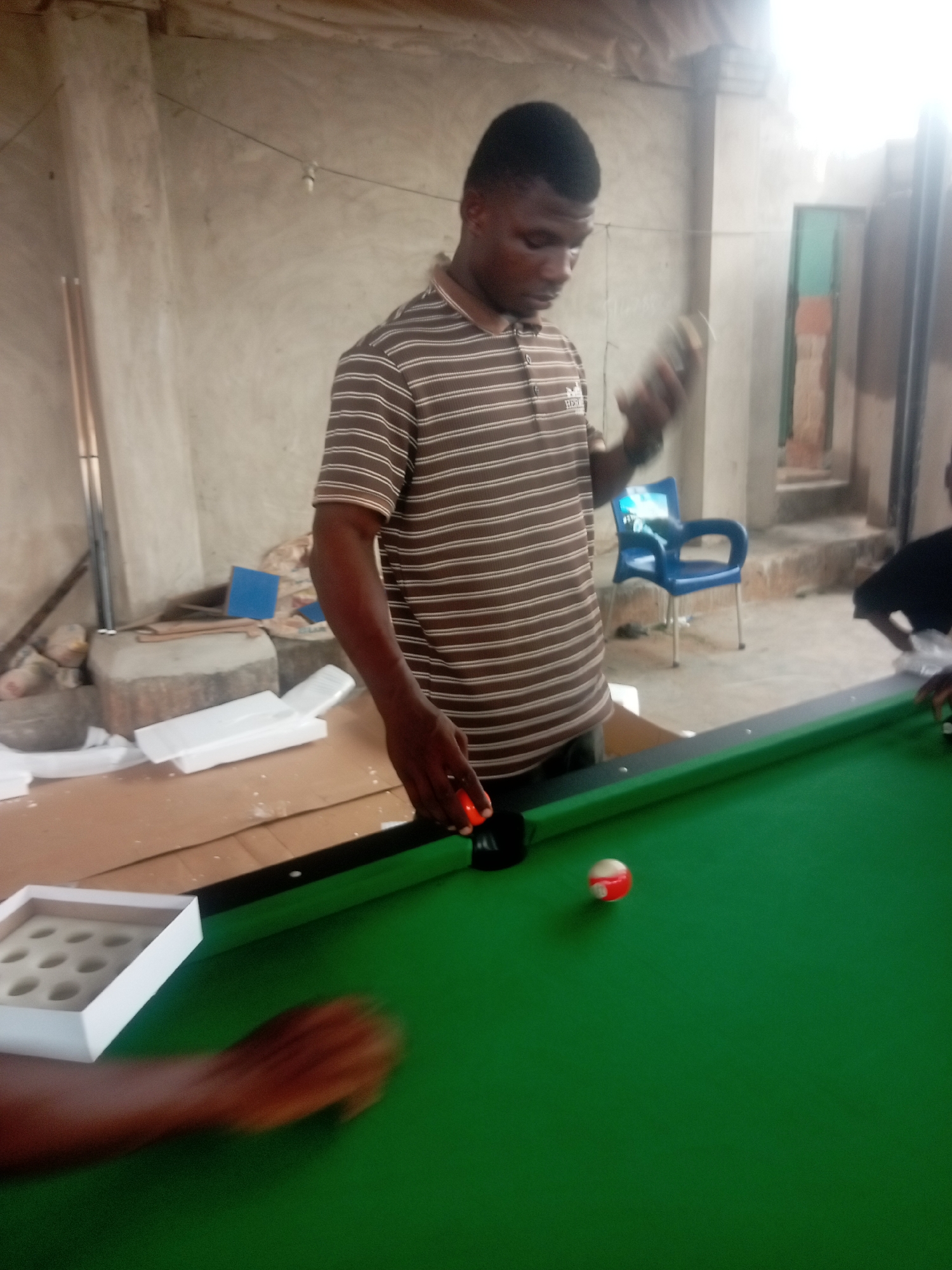 The image depicts a man standing at a green billiard table in what appears to be an indoor/outdoor space in Umuokpu, Nigeria. The man is wearing a striped brown and white shirt and is holding a billiard ball in his right hand. He is focusing on positioning a red ball on the table. Next to the table, there's a white tray with compartments, likely for holding the billiard balls. Another individual's arm, possibly holding a billiard cue, is visible on the left edge of the frame. The background suggests a simple, possibly unfinished construction, with exposed walls and a view to an outdoor area with a blue plastic chair. There's also debris and packing materials in the background, suggesting the billiard table may have been recently set up.